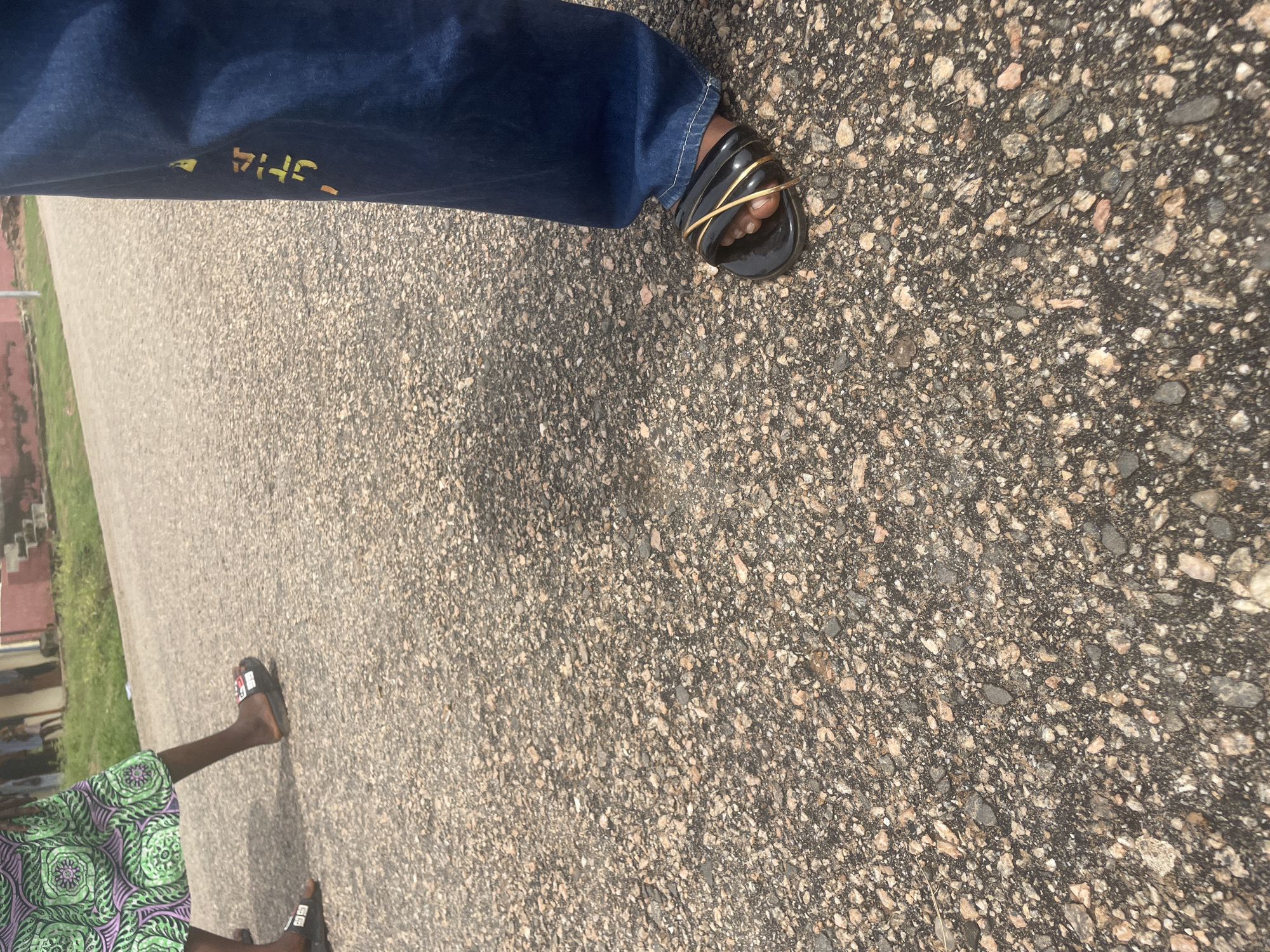 The image shows a low-angle, close-up view of two people walking on an asphalt road in Lokoja, Nigeria. The person on the left wears a green and purple patterned dress and black slip-on sandals. The person on the right wears dark blue denim jeans with a yellow and orange stencil design on the leg and black sandals with gold straps. The road surface is rough, with visible gravel and patches of discolored asphalt. In the background, there are hints of a building with a red roof and some greenery, suggesting an outdoor, possibly semi-urban environment. The lighting indicates it is daytime.