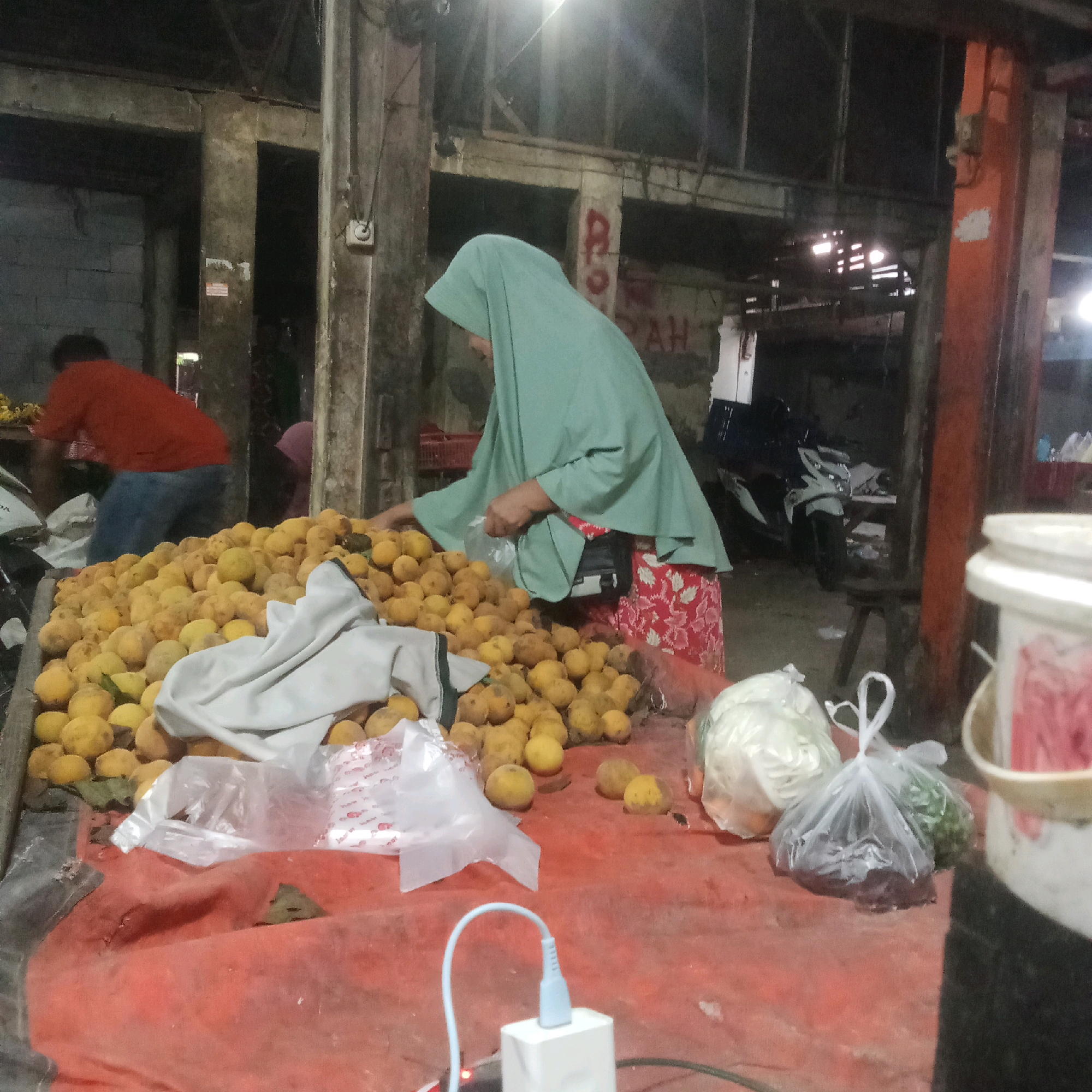 The image depicts a market scene, possibly in Karawang, Indonesia, with a focus on a stall displaying a large quantity of yellow fruits, likely limes or small mangoes, piled on a red tarp. A woman wearing a light green hijab and a floral patterned skirt is standing behind the fruit stall, reaching into a plastic bag. To her left, a man in an orange t-shirt and blue jeans is partially visible, also engaged in an activity at the stall. In the background, there are concrete pillars, a motorcycle, and various other market elements, suggesting an outdoor or semi-enclosed space. In the foreground, a white charging block with a blue cable is visible on the red tarp, indicating the presence of electronic devices. Other produce, including cabbage and what appear to be green vegetables in plastic bags, are also present on the stall. The lighting is dim, suggesting it might be evening or an indoor market.