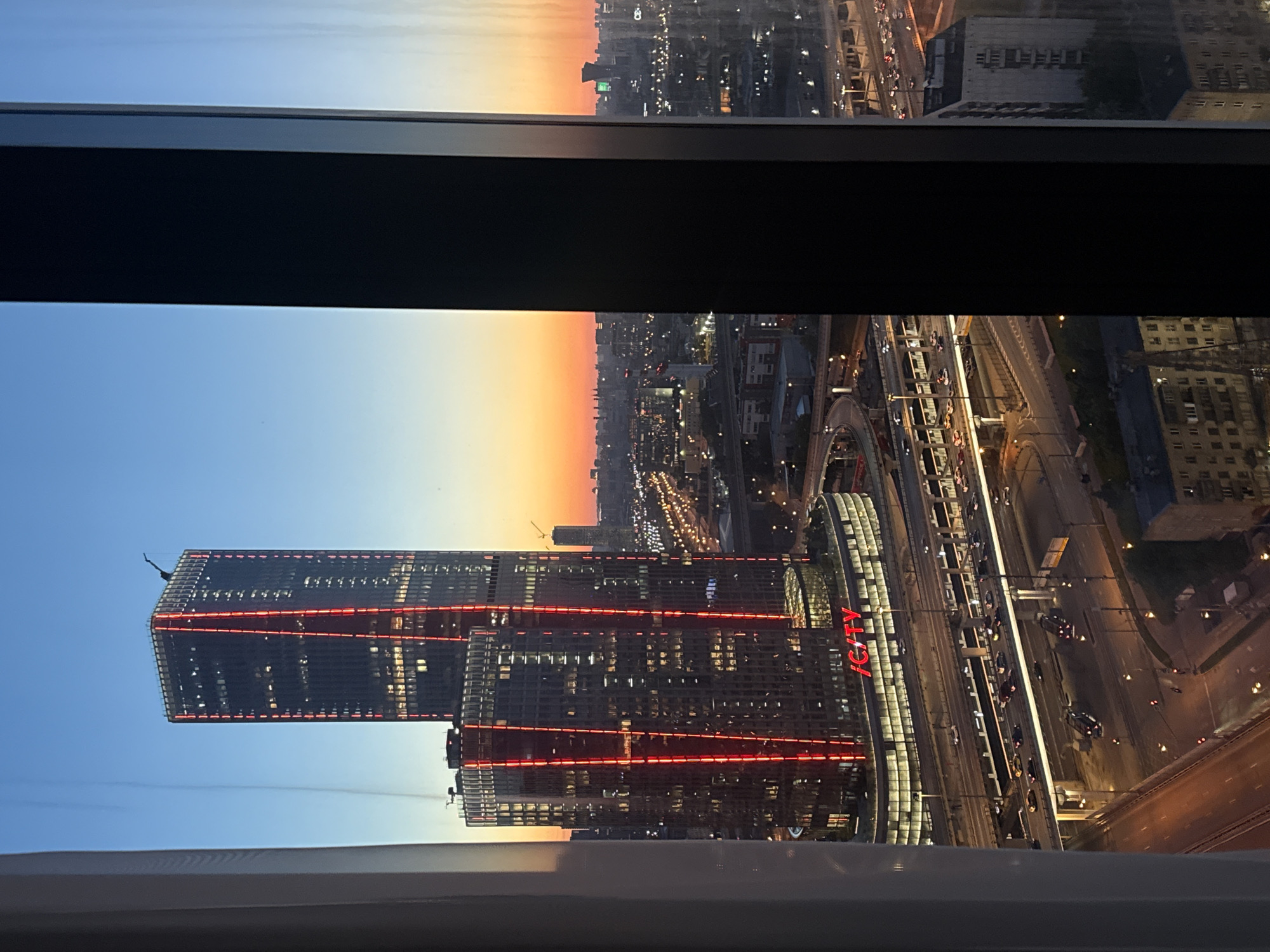 The image displays an urban landscape in Khimki, Russia, during sunset or dusk, observed from a high elevation through a window. The sky gradients from a clear light blue at the zenith to warm orange and yellow tones near the horizon. Two modern high-rise buildings with dark glass facades are prominent in the central and left portions of the view. The taller building on the far left features distinct vertical red LED strips along its edges and a silhouette of an architectural element at its top. The slightly shorter building directly in front of it also exhibits similar red LED strips configured in a "V" pattern on its visible face, and the word "iCITY" is illuminated in red text on its lower, curved structure. Numerous windows within both buildings show interior lights. Below these buildings, a complex, multi-level highway system is visible, animated by active vehicle traffic indicated by numerous car headlights and taillights, and illuminated by numerous streetlights across multiple ramps and lanes. Beyond the highway, the cityscape extends towards the horizon, showing a dense array of residential and commercial buildings with various lights on. A dark, vertical window mullion is positioned in the center-right, segmenting the view. To the right of this mullion, a continuation of the city skyline and the sunset-colored sky is visible.