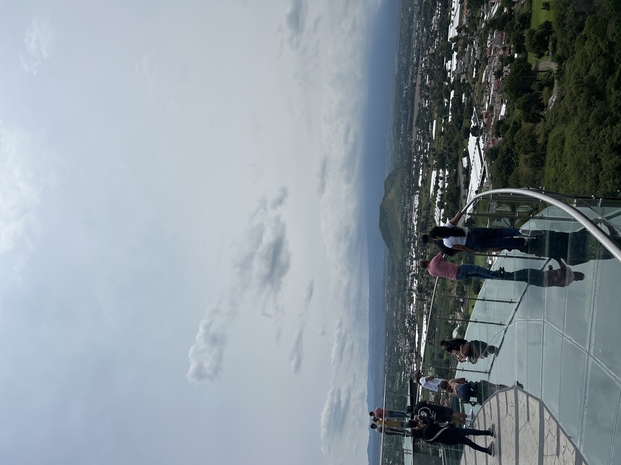 A group of people stand on a transparent glass walkway that overlooks a sprawling city and surrounding landscape under a cloudy sky. The walkway, made of glass panels, curves to the right and has a metal railing along its edge. Several individuals are positioned on the walkway, some looking out at the view, others conversing, and one is seated. The city below features numerous buildings, some with white roofs, interspersed with lush green trees and vegetation. In the distance, rolling hills and mountains are visible, partially obscured by a hazy, overcast sky with dramatic cloud formations. The foreground of the walkway shows reflections of the people and the sky on the polished glass surface. This elevated viewpoint appears to be a tourist attraction, offering a panoramic vista of the area.