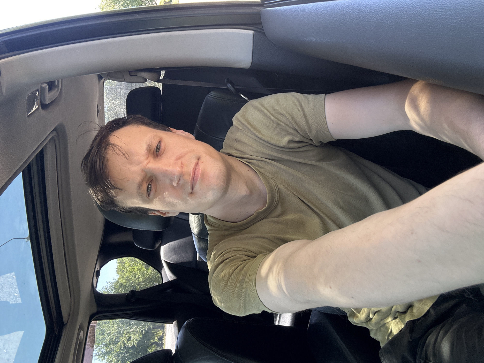 A man is seated in the driver's seat of a car, facing the camera. He has short, dark brown hair and light brown eyes. He is wearing a tan t-shirt. The car has a sunroof, and the windows are down. Outside the car, trees and a blue sky are visible. The image appears to have been taken during the daytime. The location is not specified in the image or metadata.
