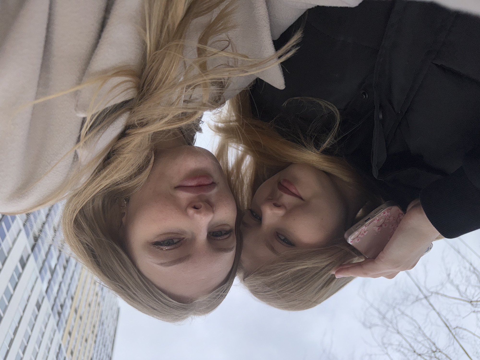 Two young fair-skinned women with blonde hair are posing closely together. The woman on the left wears a light-colored coat, while the woman on the right is in a black jacket. The latter holds a pink cell phone in her hand. A tall apartment building is partially visible behind them, and the scene appears to be outdoors during a cloudy day. The location is likely Perm, Russia.
