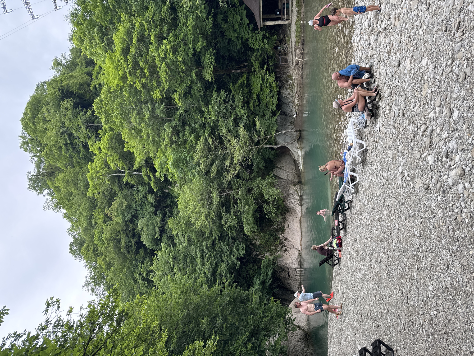 This image depicts a recreational area along a river in Khleborob, Russia. The scene is characterized by a clear, turquoise river flowing between rocky banks, with dense green forests covering the steep hillsides. On the pebble-strewn shore, several people are engaged in various activities.
In the foreground, a group of individuals are present. To the left, two men are standing, one pointing towards the water. Further down the shore, several people are seated on lounge chairs, relaxing. A few individuals are in the water, swimming or wading. One woman in a black swimsuit with a red strap is shown partially submerged in the water, with a young boy standing on the shore nearby.
On the right side of the image, a small, open-sided wooden structure with a brown roof is visible, possibly a changing room or shelter. The overall atmosphere suggests a tranquil summer day at a natural water resort.