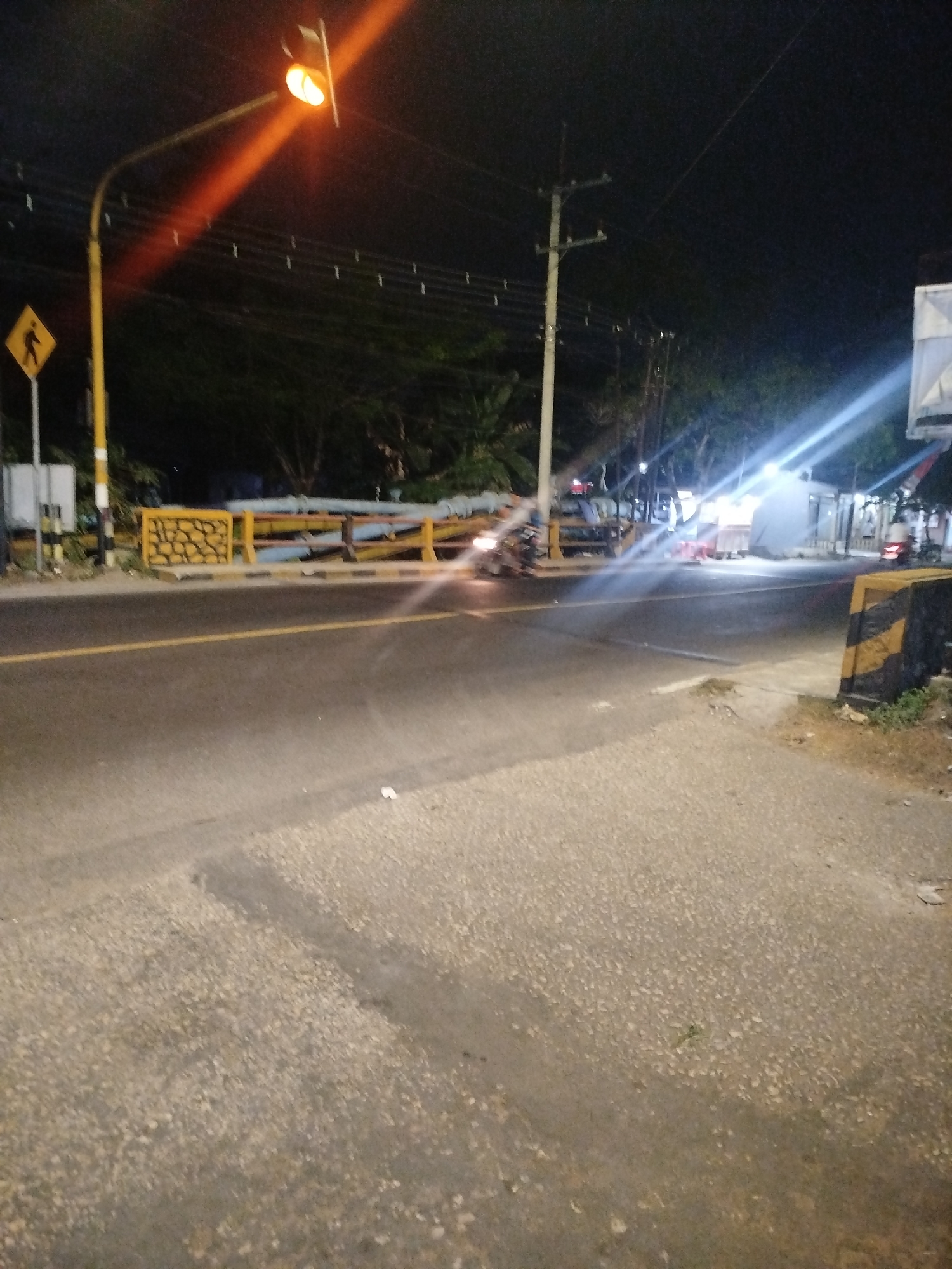 The image captures a nighttime scene at an intersection in Tonjung, Indonesia. A yellow traffic light is illuminated, casting an orange glow. To the left, a pedestrian crossing sign with a yellow diamond shape and a black pedestrian icon is visible on a pole. A yellow striped barrier runs along the side of the road. The road surface appears to be asphalt with a yellow lane marker. In the background, trees and utility poles are present. Several lights are visible in the distance, possibly from buildings or streetlights. A motorcycle with illuminated headlights is moving on the road, creating a light trail due to the motion blur. Another motorcycle is parked or stopped in the background on the right side. The ground in the foreground is a textured concrete surface.