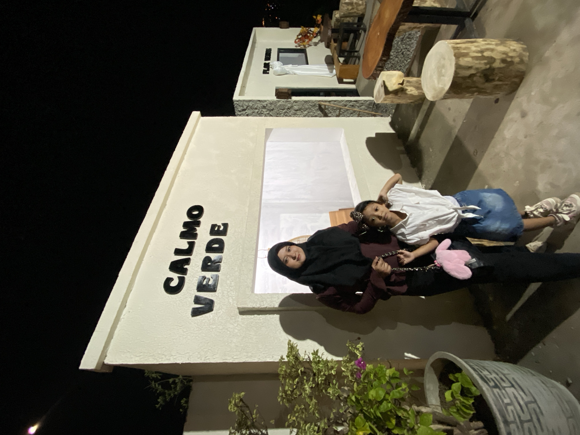 This media file shows two individuals, likely a mother and daughter, in front of a building at night. The building is light-colored and has the words "CALMO VERDE" written on it in large black letters. The woman is wearing a dark hijab and a maroon-colored top, while the child is wearing a white top and a blue skirt. They are both lying down and smiling, suggesting a relaxed and happy atmosphere.
The scene appears to be an outdoor area with a dark background, indicating it's nighttime. There are also elements of outdoor furniture visible, including a wooden table and stools made from logs, suggesting a casual dining or seating area. A large potted plant is also present in the foreground, adding a touch of nature to the scene. The overall impression is one of a peaceful evening gathering.