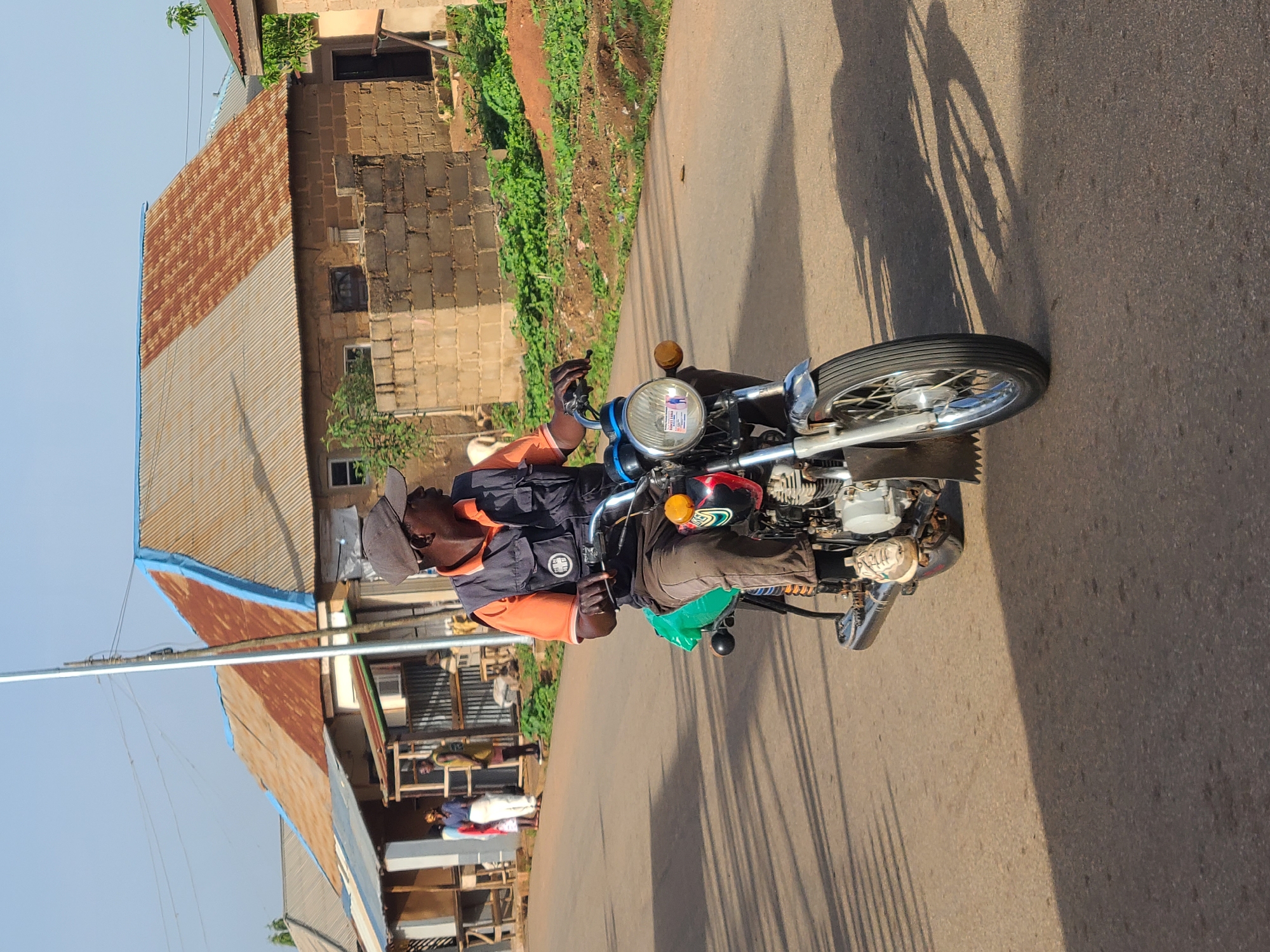 A man is riding a motorcycle on a paved road. He is wearing sunglasses, a cap, an orange shirt, and a dark vest. The motorcycle has a round headlight. In the background, there are buildings with corrugated metal roofs and unpaved walls. The location appears to be an urban environment. The location is in the city of Unknown, Unknown.