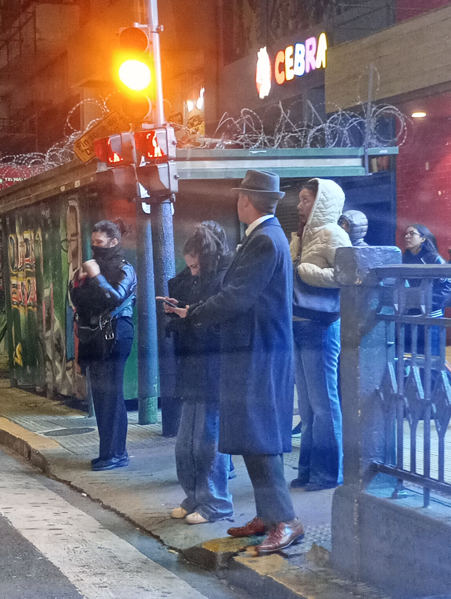 A street scene at night in Buenos Aires, Argentina, shows several people waiting at a pedestrian crossing. The traffic light is red for pedestrians. The building behind them has barbed wire on top of a flat roof above the sidewalk. A sign reads "CEBRA" in colorful letters. A woman with a mask stands to the left. A man in a long coat and hat faces away from the camera, and several other pedestrians stand nearby. A railing is visible on the right side. Graffiti is seen on the building behind the group.