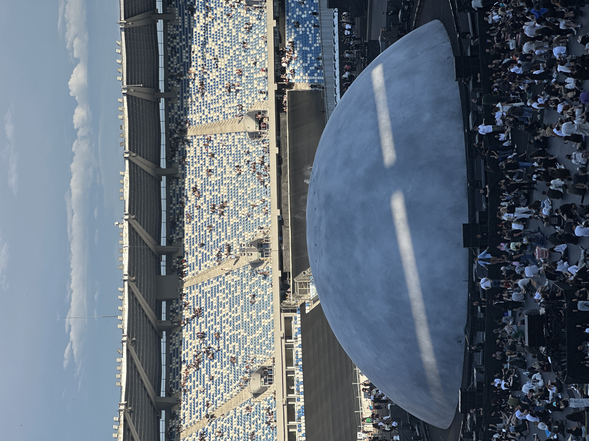The image shows a large, pale blue dome-shaped structure situated on the ground in front of a stadium with many rows of blue and white seats. Numerous people are seated on the ground in front of the dome, facing it, suggesting an event or performance. The stadium is large and appears to be outdoors. The sky above is partly cloudy with a clear blue hue. The scene is set in the Mikheil Meskhi Stadium in Tbilisi, Georgia, and the dome is a stage setup for a concert.