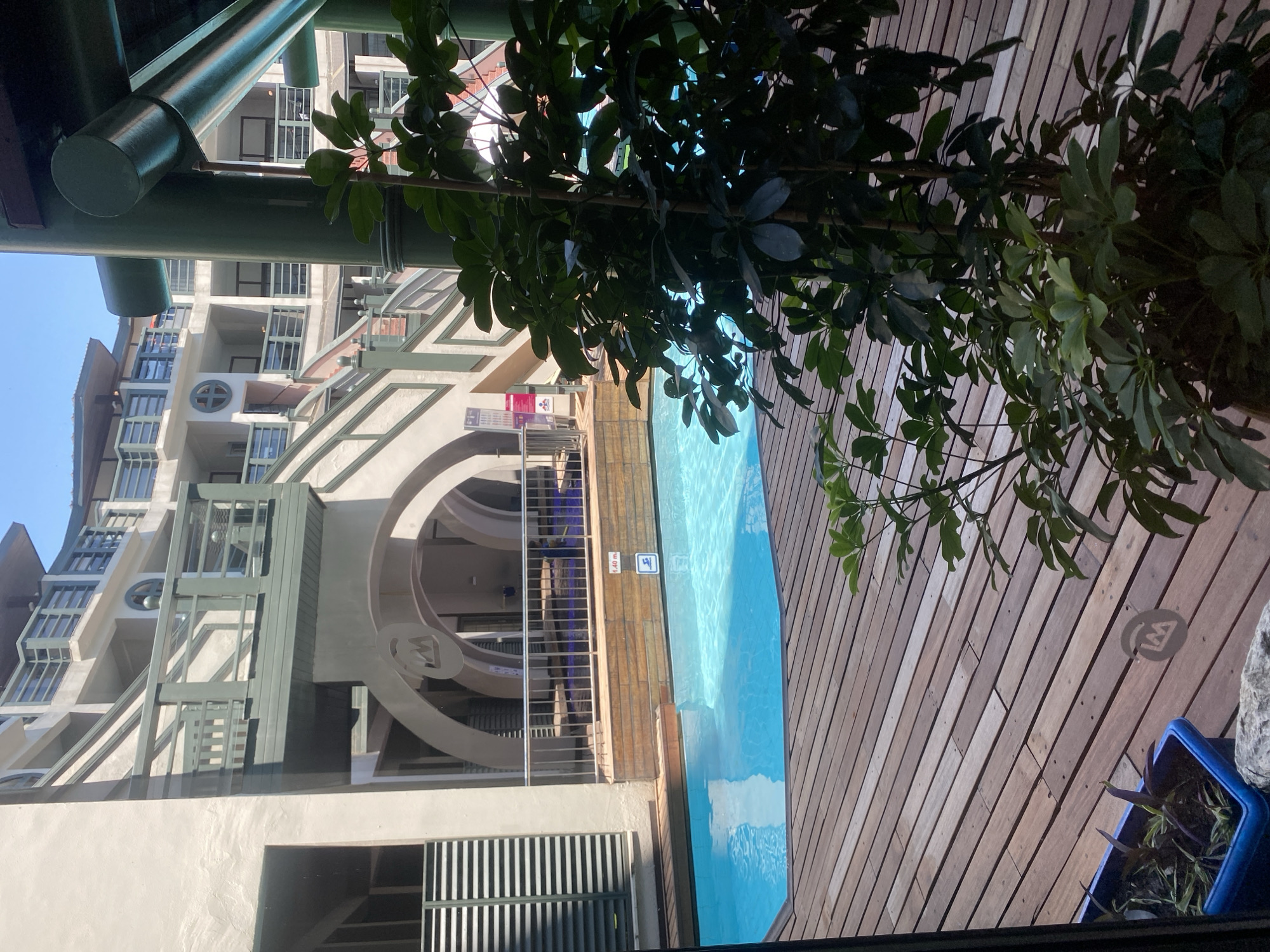 The image shows an outdoor swimming pool area with a wooden deck. In the foreground, there are potted plants. The pool is surrounded by a wooden deck. In the background, there is a multi-story building with balconies and staircases. The architecture is modern with a focus on geometric shapes and curved lines. There are no people visible in the image. The location is identified as Aksu, Turkey.