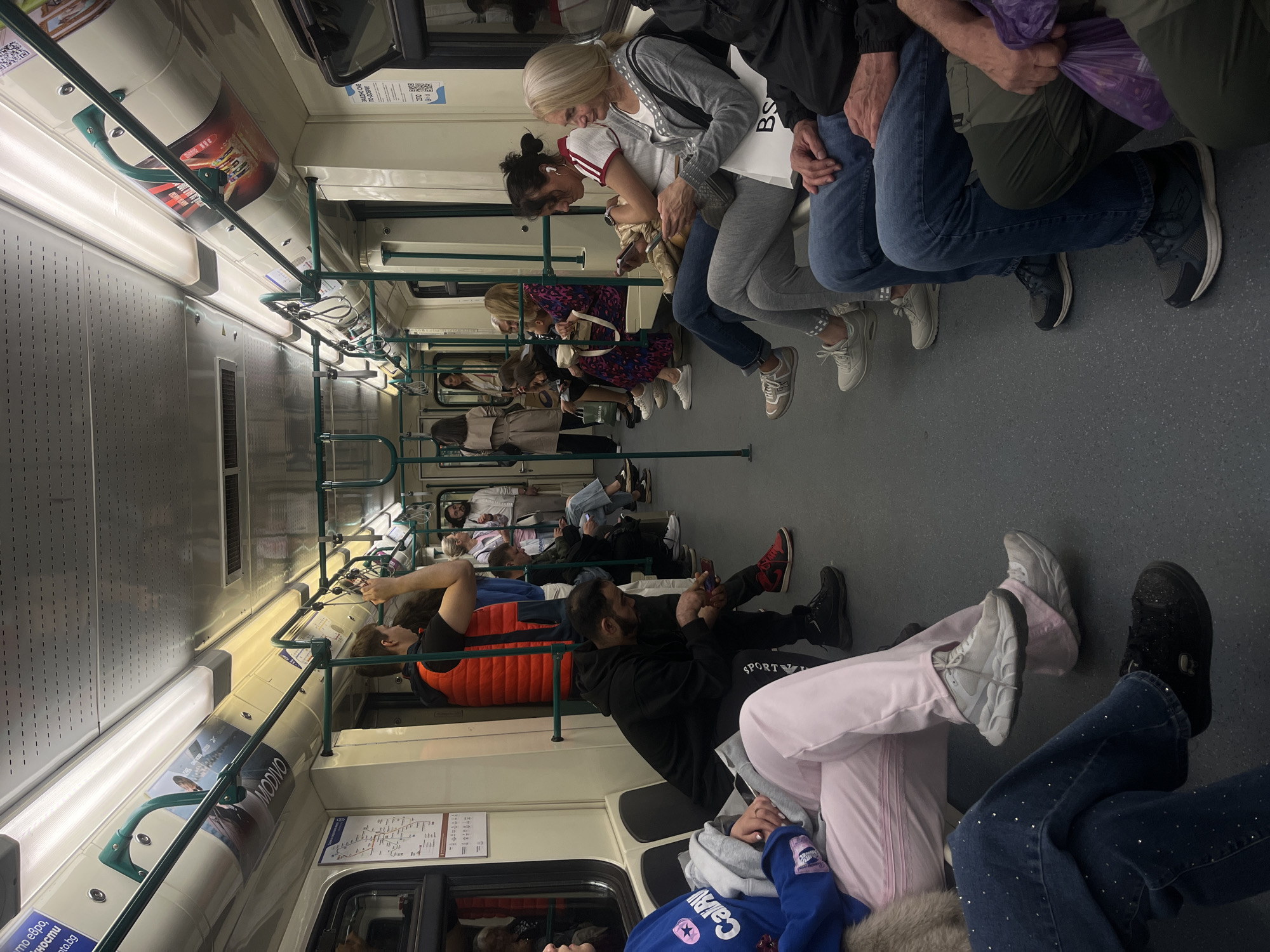 The image captures the interior of a public transport vehicle, potentially a subway or train, populated with numerous passengers. Many are seated, while others are standing, utilizing the overhead handrails for support. The interior of the train car features advertisements and informational signage affixed to the walls. The carriage is well-lit by overhead fluorescent lighting. The scene is characterized by the close proximity of passengers and the generally quiet, orderly atmosphere typical of public transit.