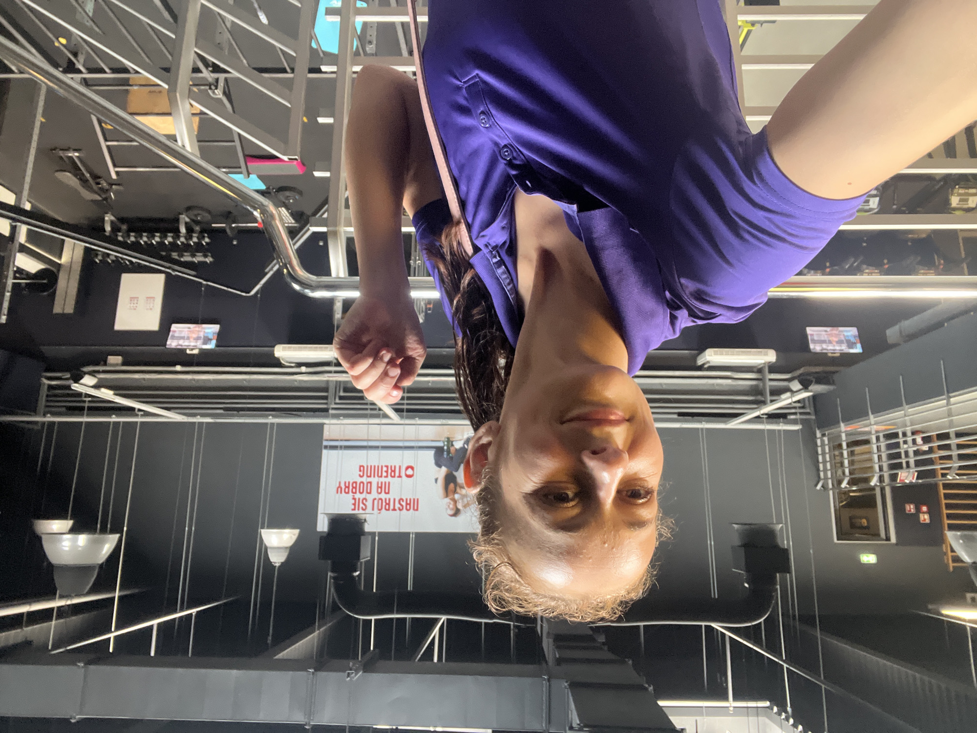A woman is taking a selfie in a gym. She is wearing a purple polo shirt. The gym appears to be multi-level with exercise equipment visible in the background, including treadmills and weight racks. There is also a large banner on the wall with Polish text and images of people exercising. The lighting suggests it is indoors.