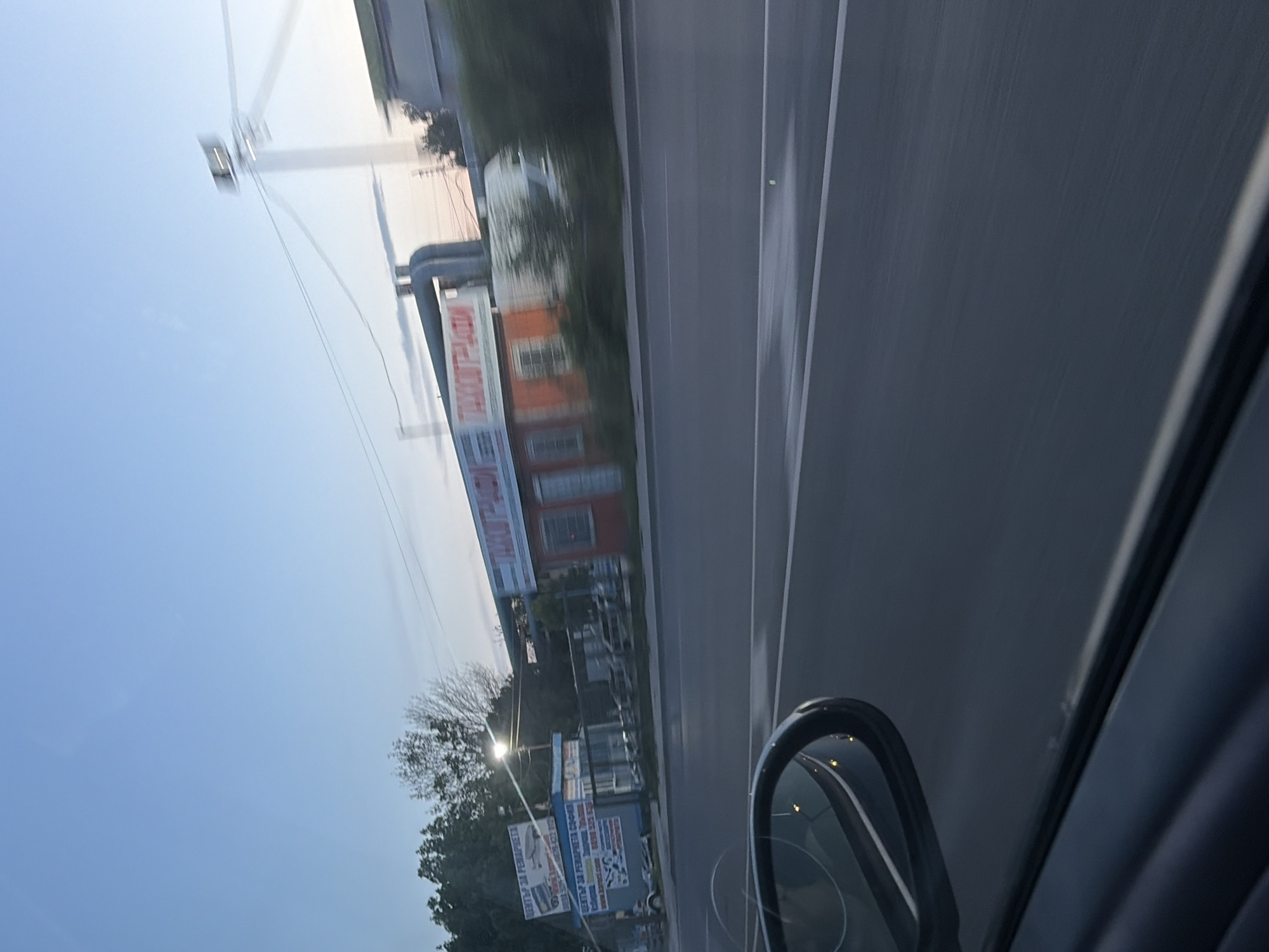 The image depicts a view from inside a car driving along a road in Sofia, Bulgaria. On the left side of the road is a building with a sign in Cyrillic script, followed by trees, streetlights, and additional signage. An electrical tower is also visible. The road itself is asphalt with white lane markings. Part of the car's interior is visible on the right. The sky is visible above the buildings and trees.