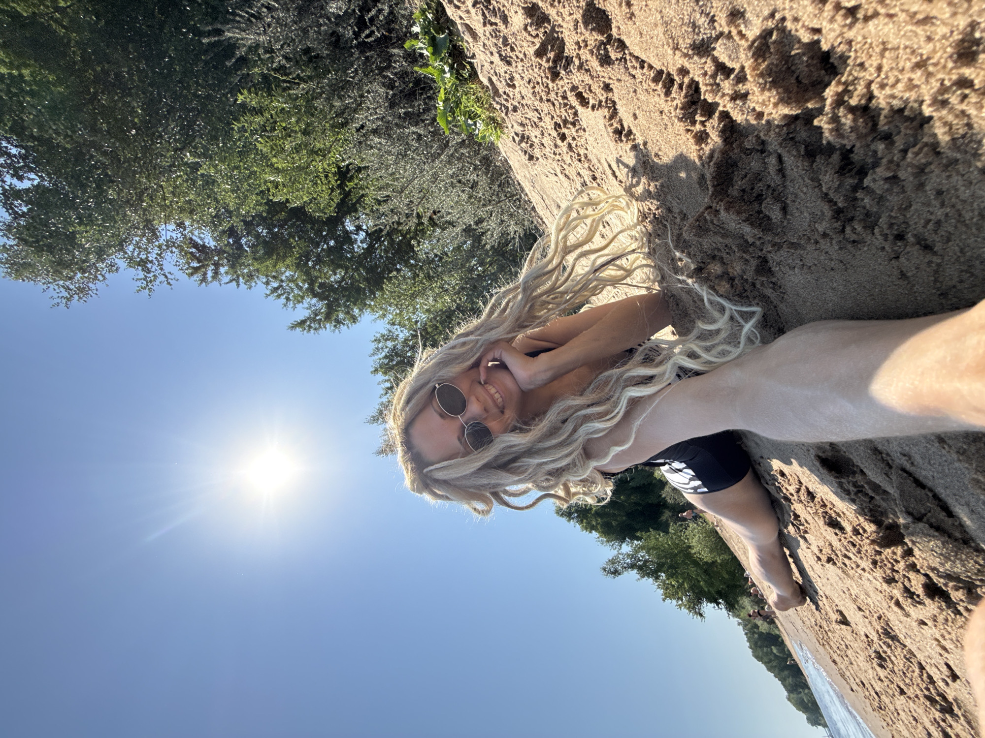 A woman with long, blonde, dreadlocked hair and round sunglasses is lying on a sandy beach, possibly taking a selfie. She is wearing a black swimsuit. The sun is visible in the upper left corner of the image, shining brightly against a clear blue sky. Trees are visible at the top and bottom of the frame. The location is implied to be Rusanovo, Russia. The woman is smiling and has one hand near her face. Water is at the bottom edge of the frame.