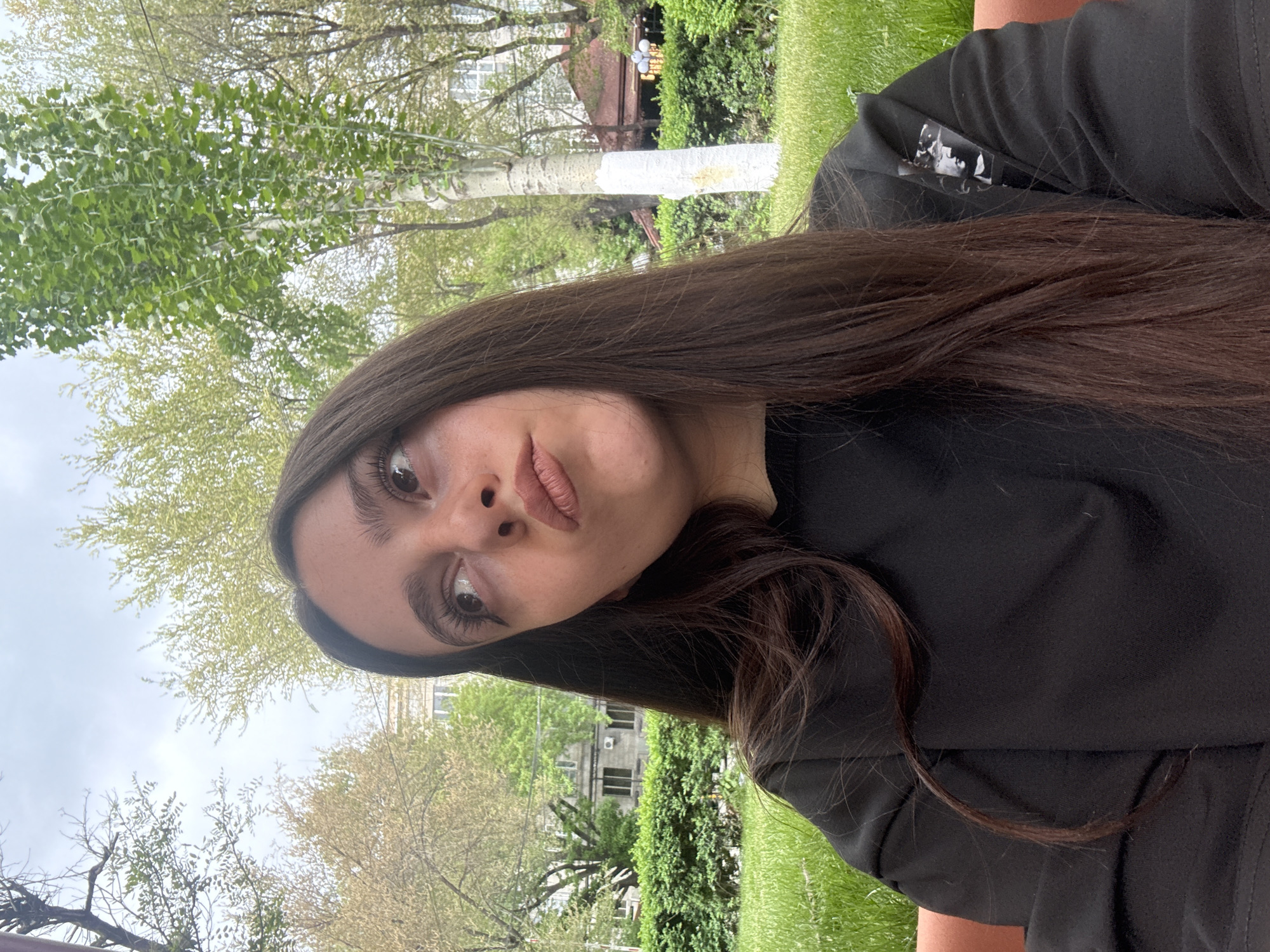 A woman with long, dark brown hair is the central subject of this selfie. She has brown eyes, fair skin, and pursed lips. She is wearing a black shirt.
The background features an outdoor setting with grass and trees in the daytime. A building is visible in the distance, and a white tree trunk is also prominent.
Based on the architecture and foliage, this image was likely taken in the city of Yerevan, Armenia.