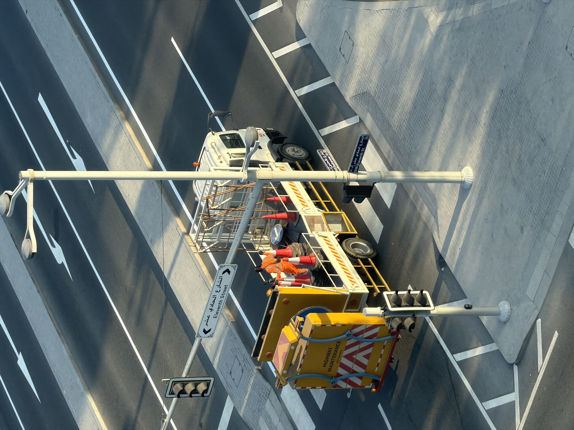 An aerial view shows a highway maintenance truck in the city of Unknown, Unknown. The truck, which is white with a yellow and red service box on the back, is stopped at a pedestrian crossing on a street. A worker in an orange high-visibility jumpsuit is standing on the back of the truck, near several orange traffic cones. The truck has "HIGHWAY MAINTENANCE" written on the back. The street has white lane markings and directional arrows painted on the asphalt. There are traffic lights and street signs visible. The scene is lit by sunlight, casting shadows across the road and sidewalk.