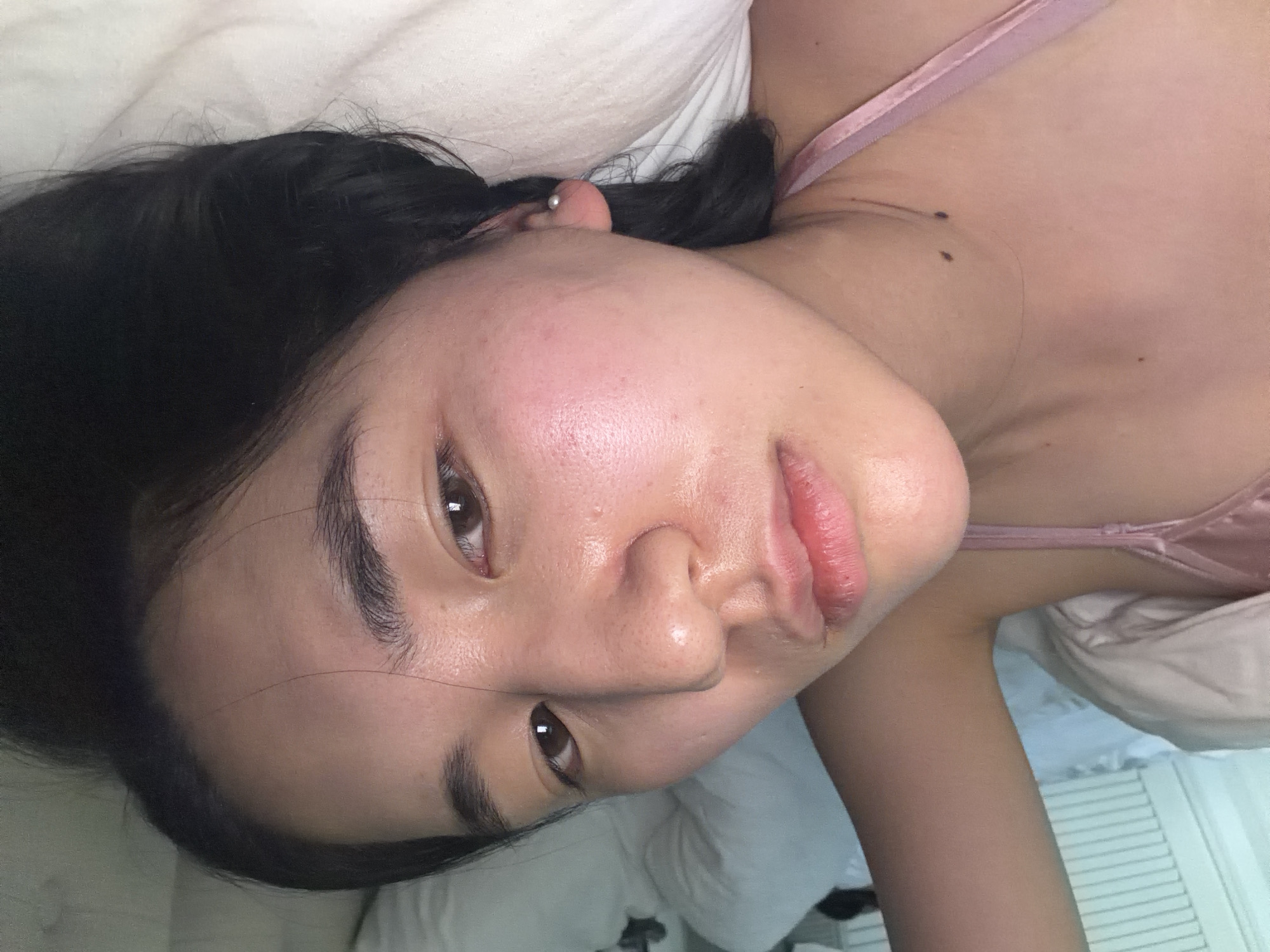 A young woman with dark hair is lying in a bed with white sheets and a light pink satin camisole. She has fair skin and is looking directly at the camera. The background appears to be a bedroom with white walls. There is no information in the provided image to suggest that this image was taken in the city of Grimaud, France.