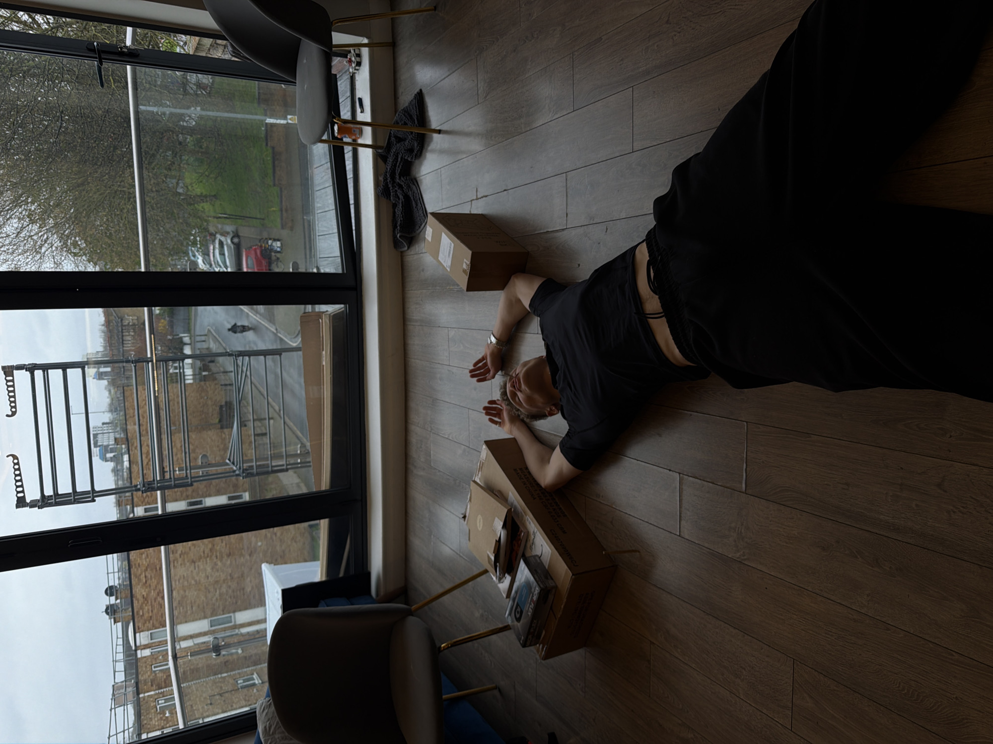 The image depicts an indoor scene within a modern apartment, viewed through large glass balcony doors opening onto an urban street in London, United Kingdom. An adult individual lies supine on a light brown wooden floor in the foreground, wearing a black short-sleeved top with a visible midriff and black loose-fitting trousers. Their arms are bent, with hands resting on their forehead; a silver watch is present on the left wrist.
To the individual's left, a stack of cardboard boxes rests on the floor. The bottom box bears text including "MADE IN CHINA." On top of this are a smaller, flatter cardboard package, a black retail package labeled "MDR FLEX," and several compact discs. A separate, smaller brown cardboard box is positioned to the individual's right.
Two modern dark grey chairs with gold-colored metal legs are visible near the window frame. A blue upholstered furniture item is partially visible behind the left chair. On the right window ledge, an orange soda can, a small black object, and a small white object are present. A dark grey towel or cloth is crumpled on the floor near the right chair. A metallic multi-tier drying rack is propped vertically against the left glass panel of the balcony door.
Outside, the street appears wet under an overcast sky. The view reveals multi-story brick buildings, mature trees with bare branches, parked vehicles including red and silver cars, and a single individual operating a moped on the street. Scaffolding is affixed to a building in the distant background.