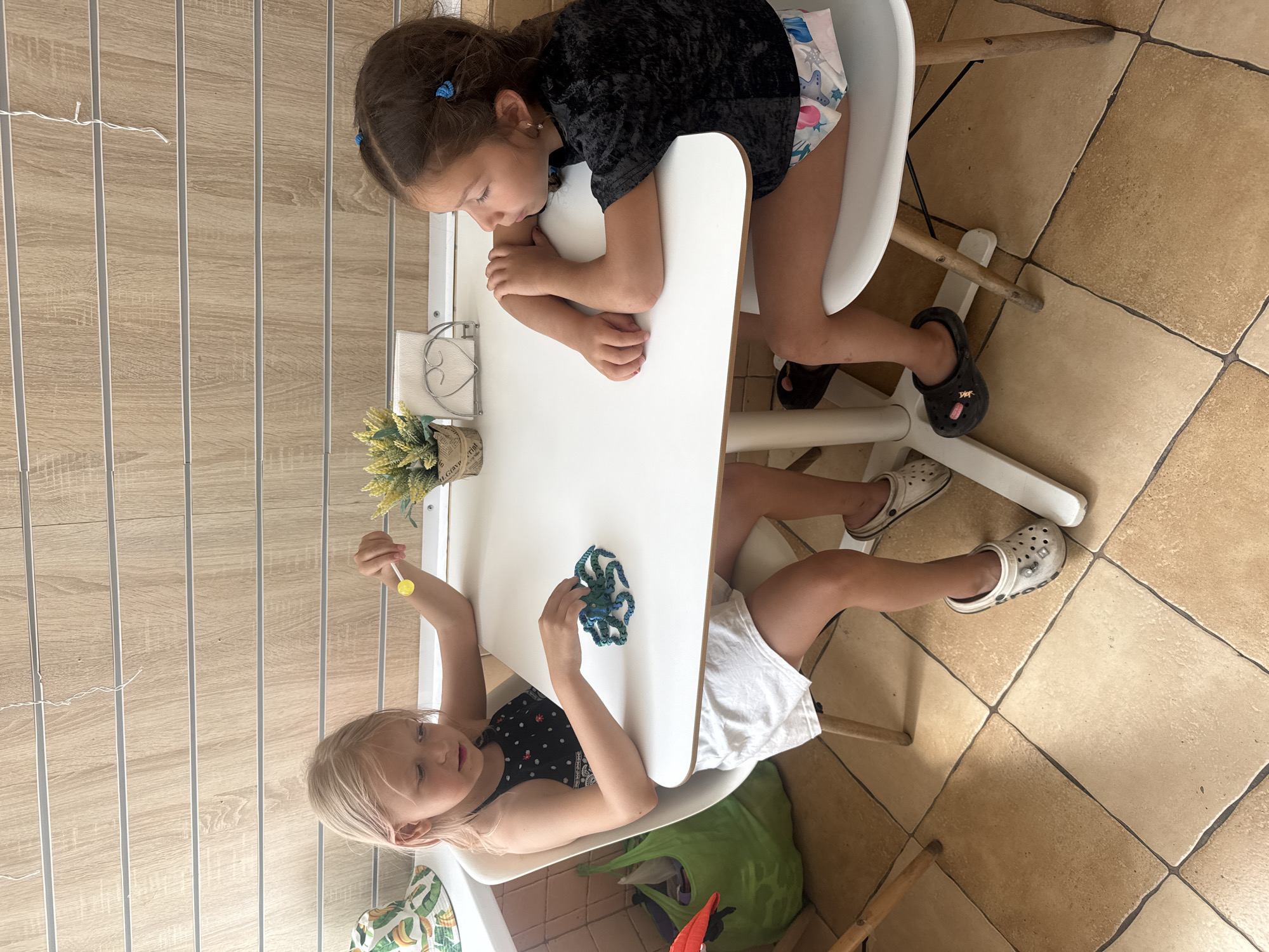Two young girls are seated at a white table, facing each other. The girl on the left, with blonde hair, is wearing a black and white patterned top and white shorts. She is holding a yellow lollipop in her right hand and a green, octopus-like toy in her left hand on the table. The girl on the right, with brown hair, is wearing a black top and patterned shorts. She has her arms crossed and is looking down at the table. A plant in a pot and a napkin holder are visible on the table between them. The setting appears to be indoors, with wooden-paneled walls in the background and a tiled floor. A green bag is on the floor to the left of the table. The scene does not provide specific details that would allow for identification of the location as Partenit, Ukraine.