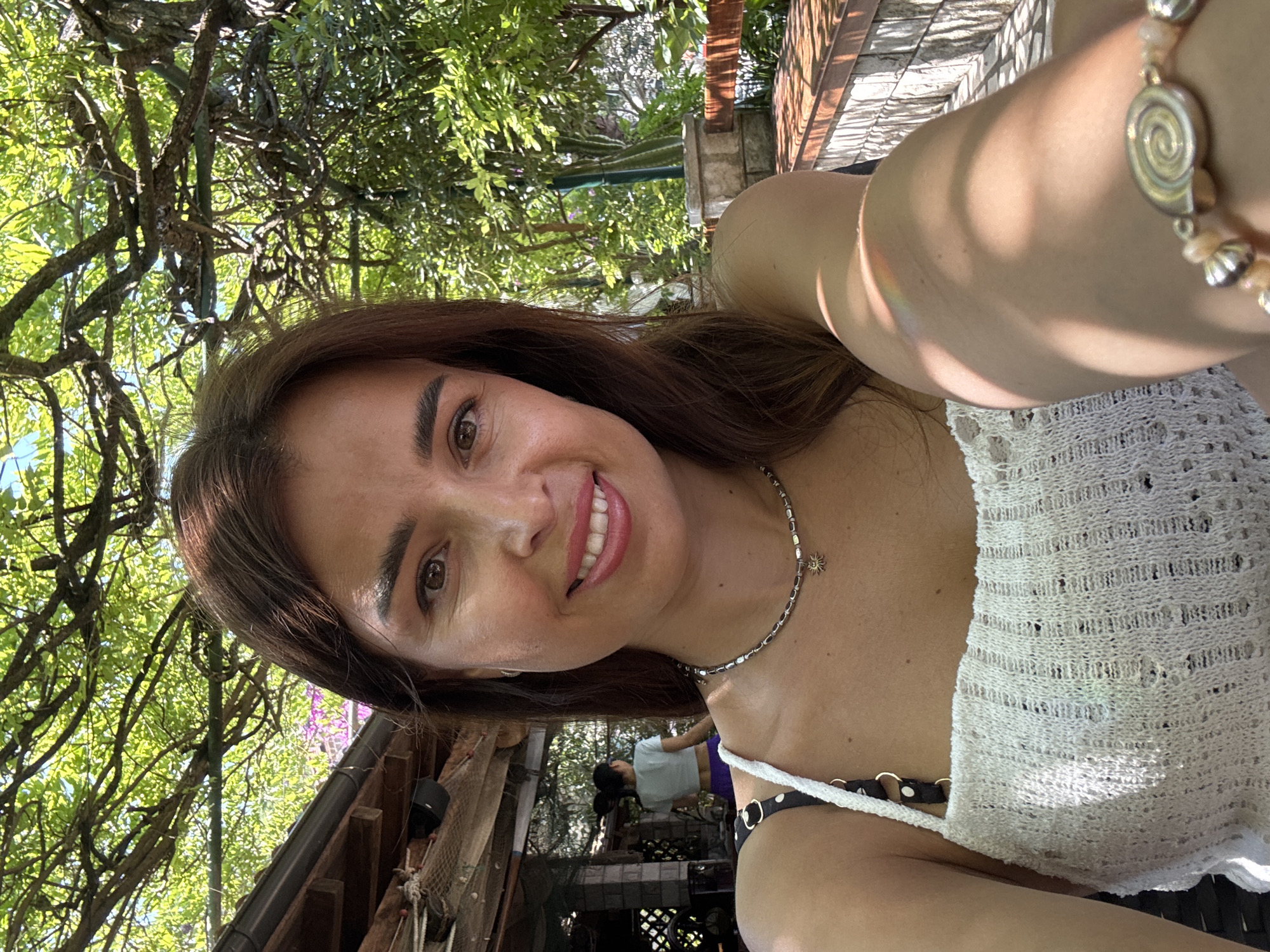A woman with brown hair and brown eyes smiles at the camera in a selfie. She is wearing a white, crocheted tank top with black polka dot straps. Around her neck is a silver necklace with a sun pendant. She has a bracelet on her right arm with a spiral design. The background is a garden setting with dappled sunlight filtering through green tree branches. In the background, another woman with her hair in a bun is visible, wearing a light blue shirt and purple pants. There is a wooden awning and some netting on the left side of the image.