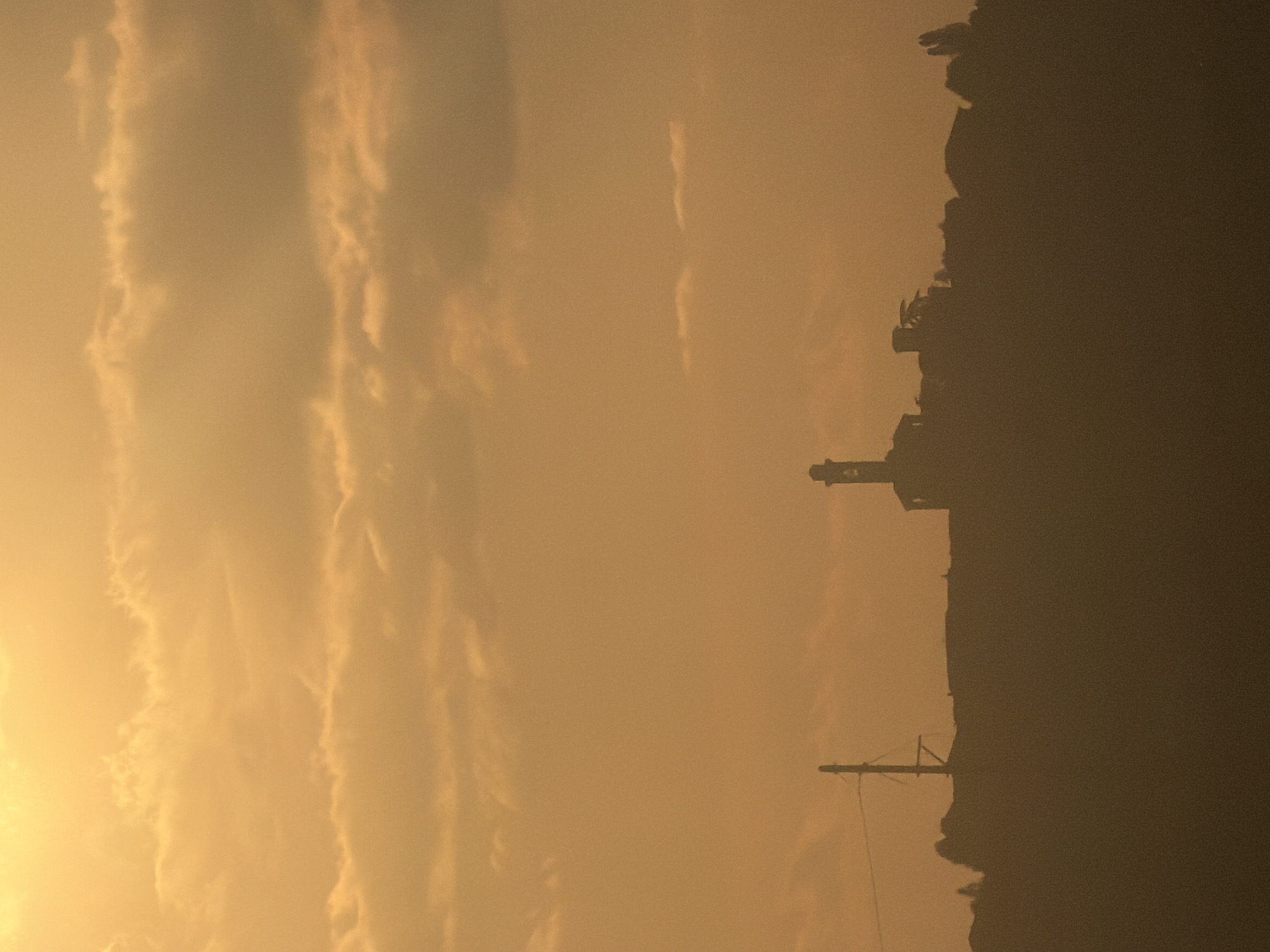 The image depicts an orange sky at sunset in Santo Stefano Belbo, Italy. Silhouetted against the sky are a line of trees, a utility pole, and what appears to be a tower. The sky is filled with long, streaky clouds.