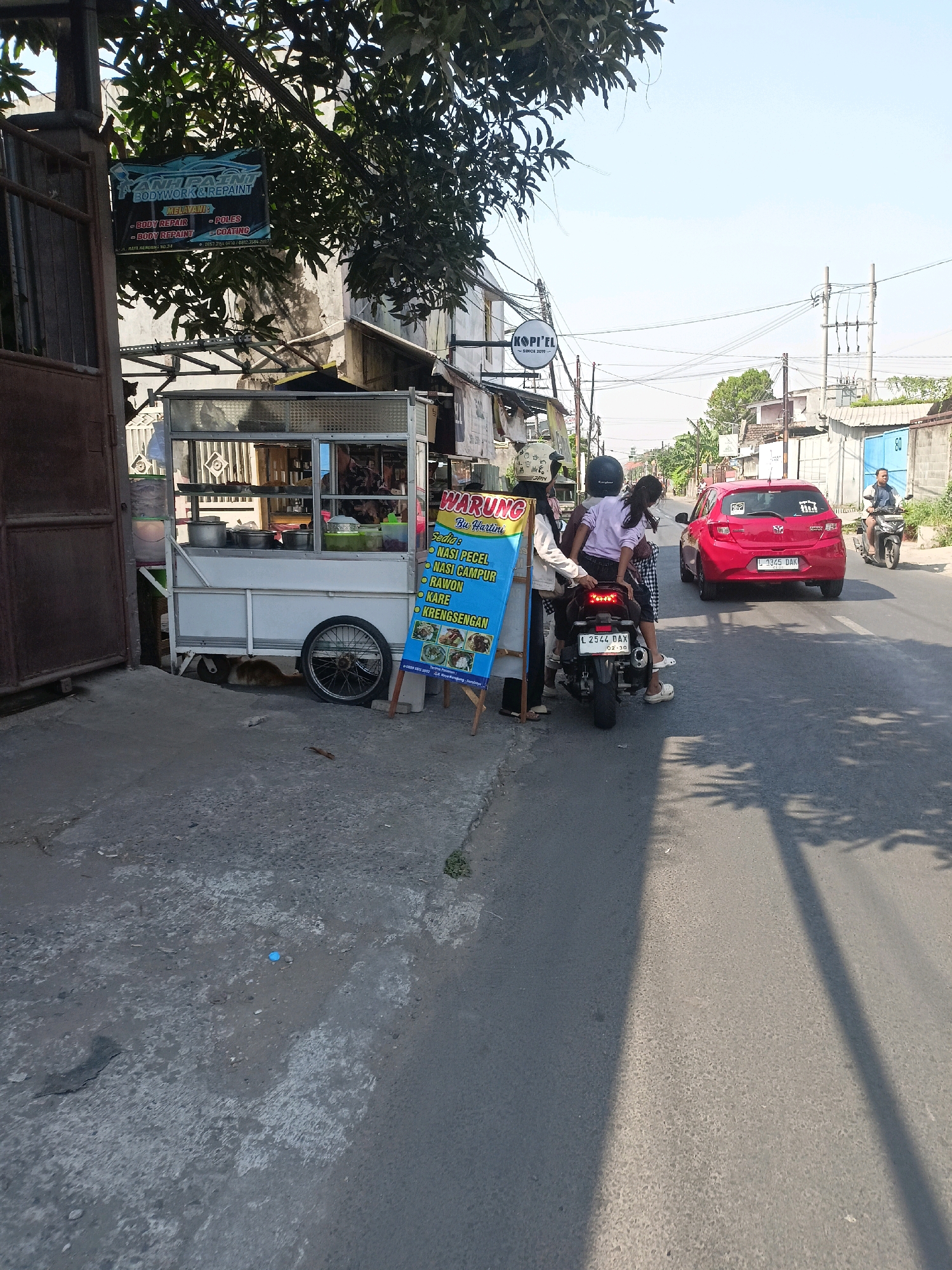 In Surabaya, Indonesia, a street scene unfolds under a bright sky. A food cart, known locally as a "warung," stands on the left side of the frame, its white structure partially obscured by a leafy tree. The cart displays a menu board advertising Indonesian dishes such as "Nasi Pecel," "Nasi Campur," "Rawon," "Kare," and "Kareng Sengan." A red hatchback car, with license plate L 1345 DAK, is parked on the right side of the street, facing away from the viewer. A man on a motorcycle, wearing a blue shirt, rides past the car in the distance. In the foreground, a group of individuals are gathered around a motorcycle. One person, wearing a helmet and a white jacket, stands next to the motorcycle, while another, dressed in a purple top and patterned skirt, sits behind the rider. The street is paved with asphalt and shows signs of wear and tear. The environment is urban, with buildings and power lines visible in the background.