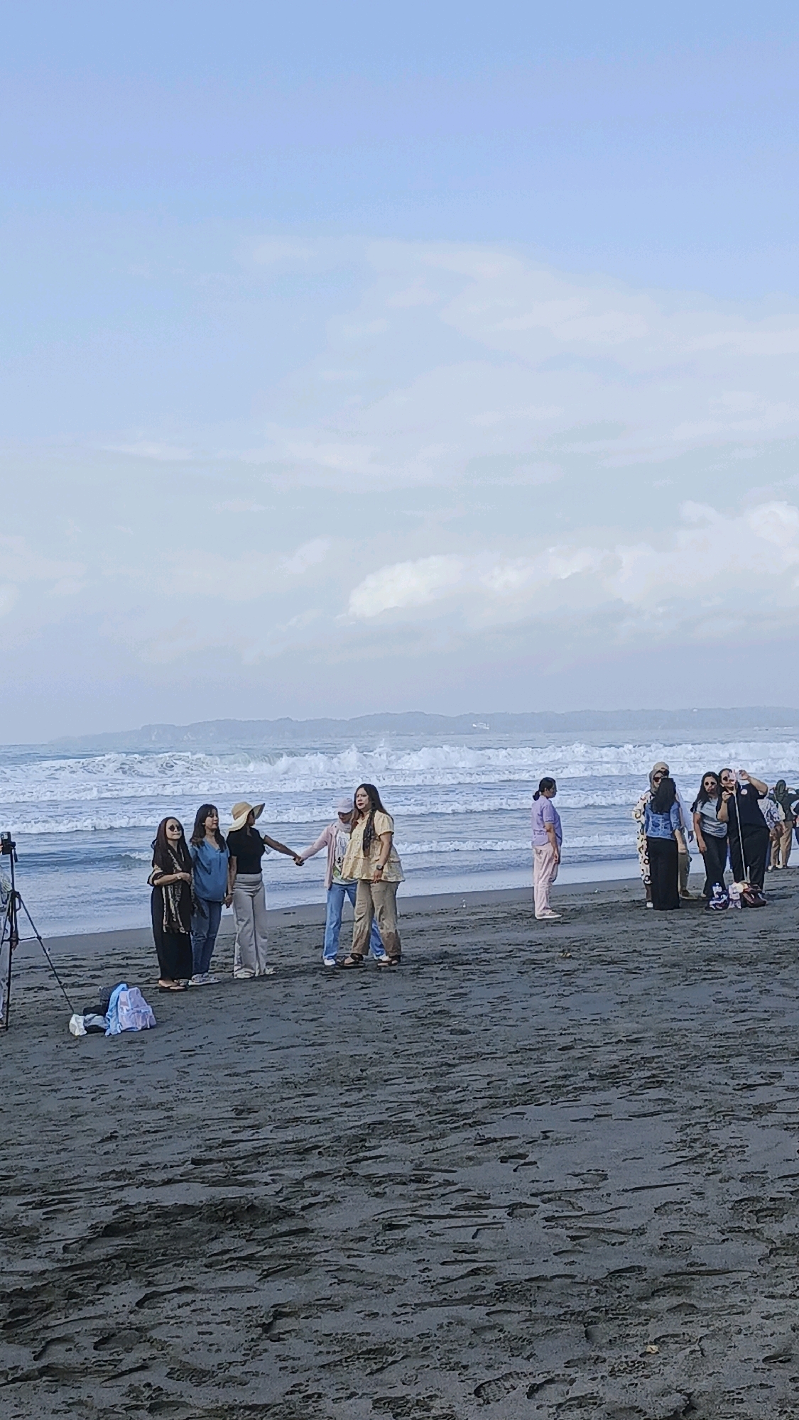 In the city of Desa Ciliang, Indonesia, a group of eight women are gathered on a dark sand beach with the ocean in the background. Five women are in the foreground, with four of them holding hands in a line. They are dressed in various casual outfits including pants, skirts, and tops. One woman in the foreground is wearing a straw hat and a black top with grey pants. To their right, a group of three women stand together, with one appearing to take a photo with her phone. The ocean is moderately wavy with a distant, hazy shoreline and mountains. The sky is overcast with light grey clouds. A tripod for a camera is visible on the left side of the image, and a light blue bag is on the sand near it.