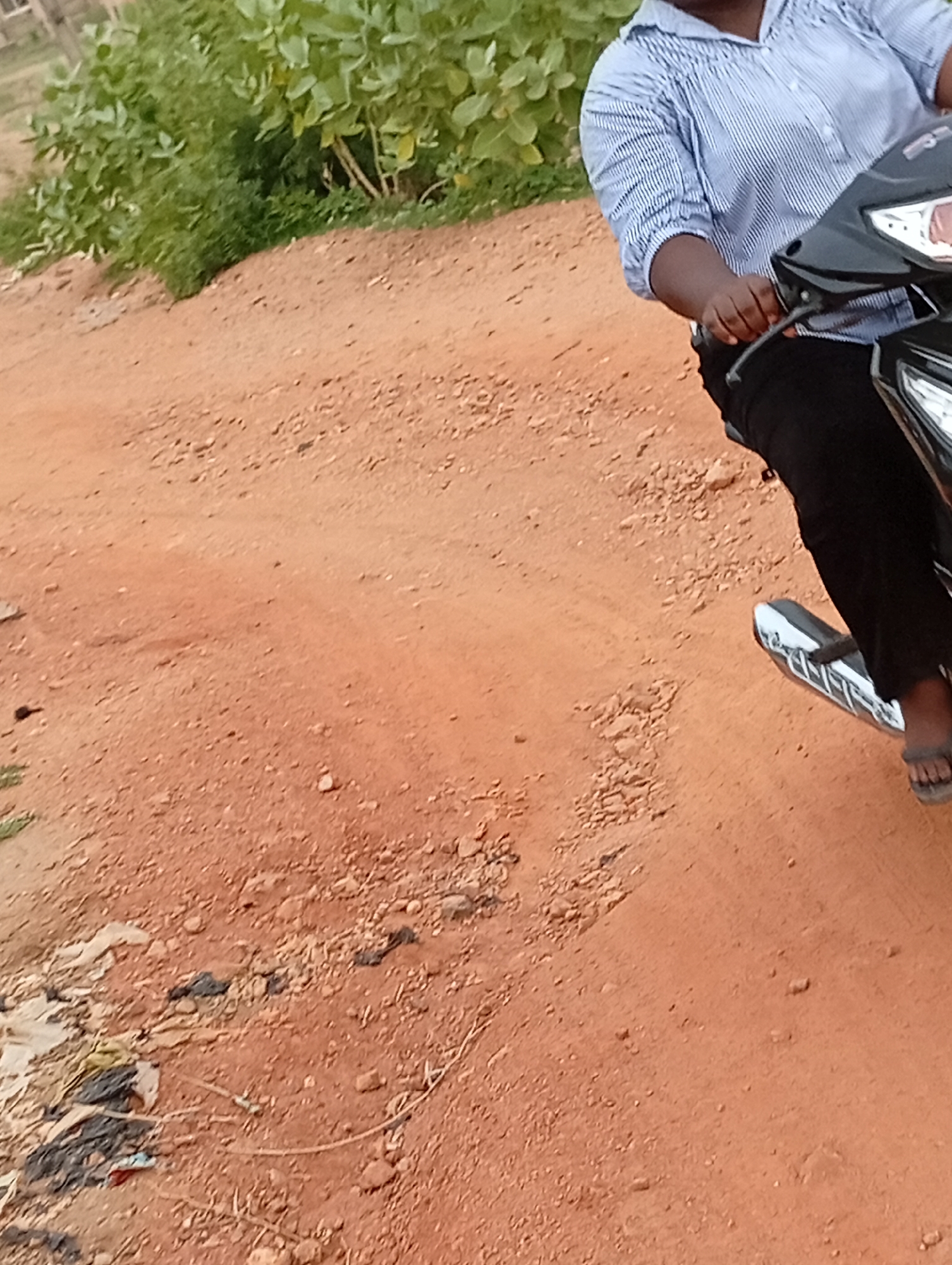 A medium-shot image shows a person riding a black scooter on a reddish-brown dirt road in what appears to be the city of Adankolo, Nigeria. The person is wearing a blue and white striped shirt and black pants, with open-toed sandals. The surroundings include green vegetation on the left side of the image. Debris litters the ground.