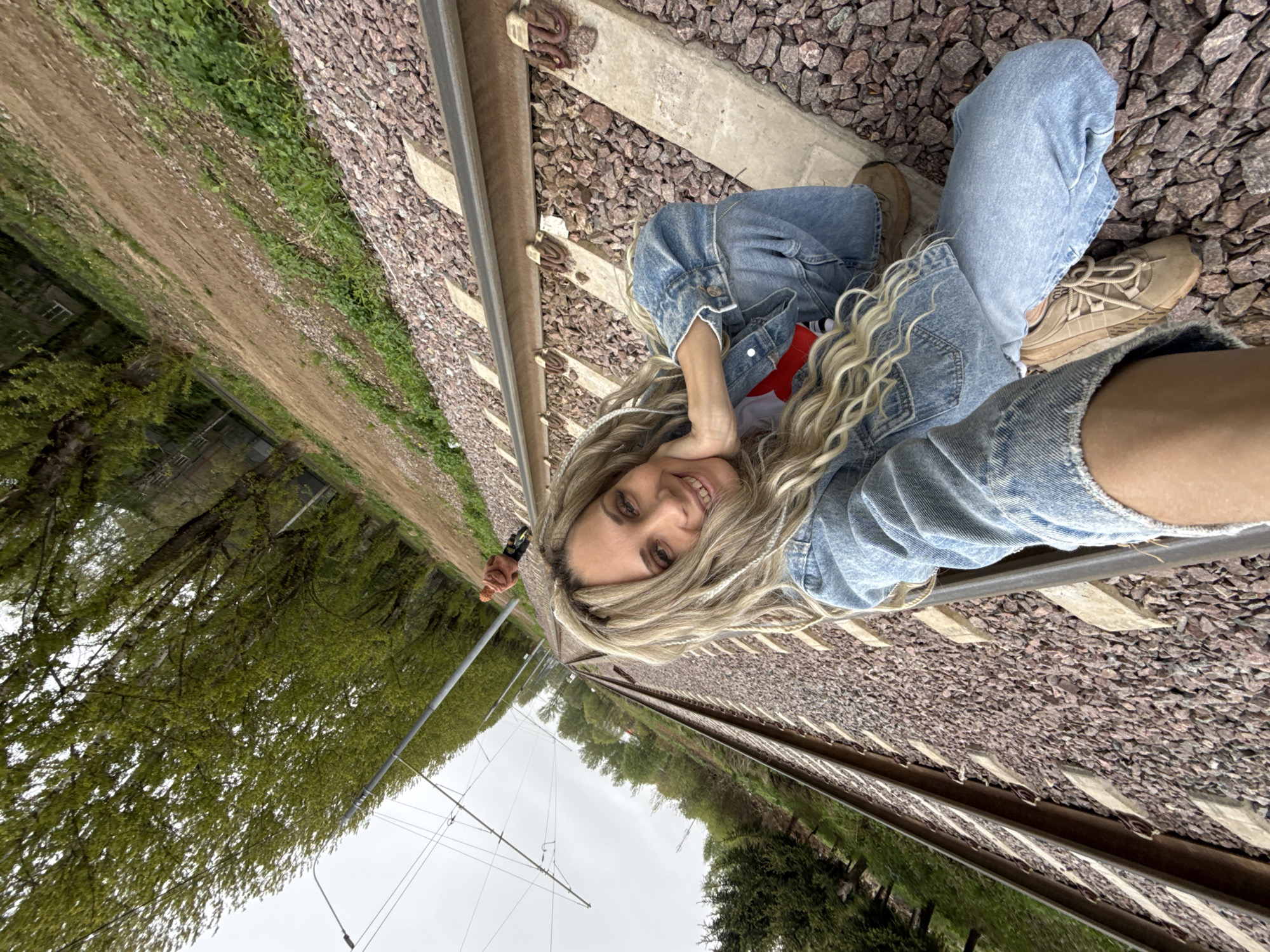 A woman is posing for a selfie while lying on a set of railroad tracks in Rusanovo, Russia. She is wearing a denim jacket, jeans, and sneakers. The tracks are surrounded by gravel and wooden sleepers. Trees and a dirt path line the edge of the tracks. Overhead power lines and utility poles are visible. In the background, another person is visible on the pathway. The sky is overcast.