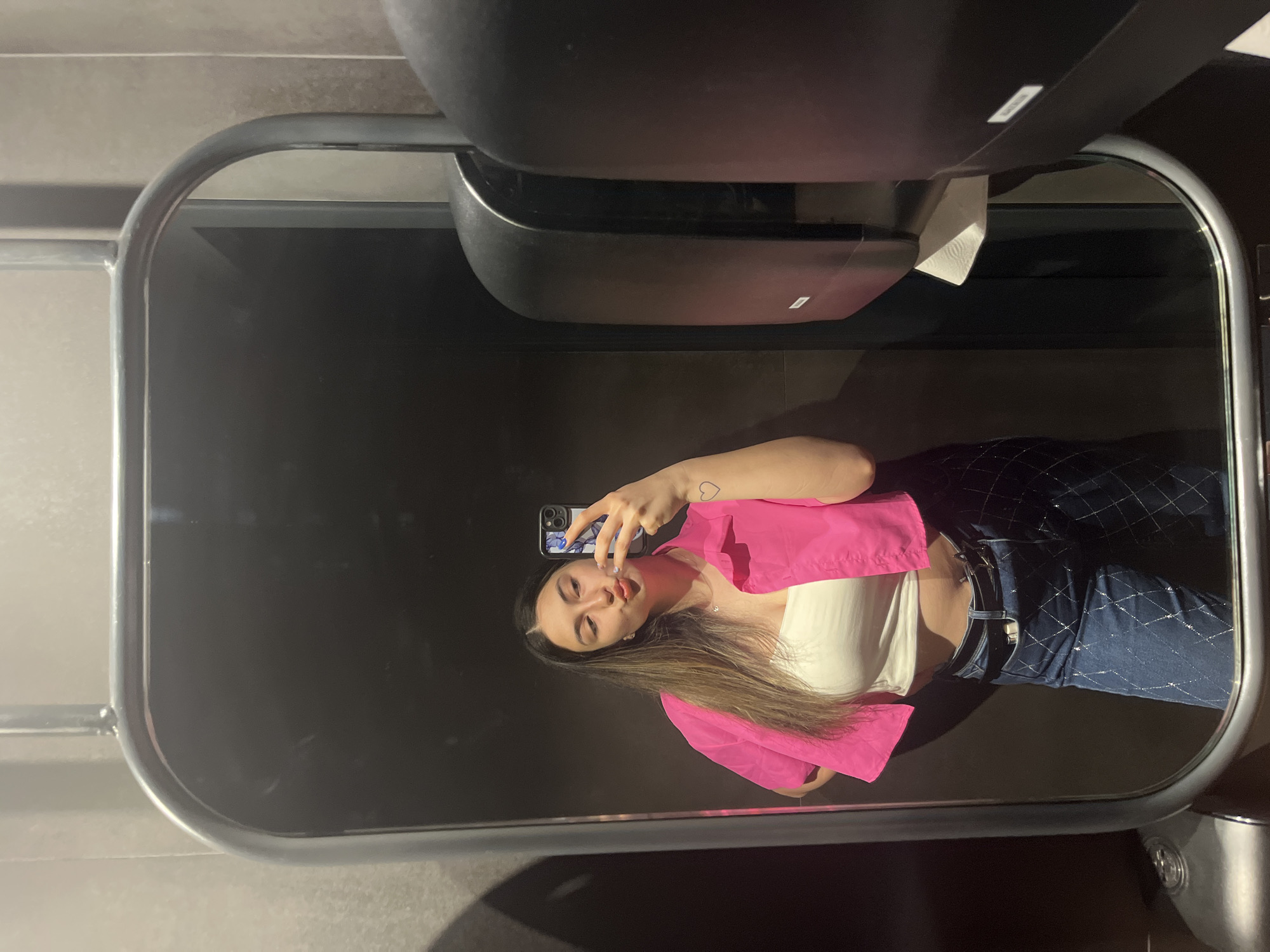 A young woman is taking a mirror selfie in what appears to be a public restroom. She is wearing a bright pink crop top over a white bustier and blue jeans with a diamond pattern. She has long brown hair and is making a kissing face, holding her phone up to take the photo. A small heart tattoo is visible on her left forearm. To the right of the mirror, two black dispensers are mounted on the wall, one of which has paper towels hanging out. The environment is dim with a tiled wall behind the woman and a darker tiled wall on the right. The location is not identifiable as being in Plovdiv, Bulgaria.