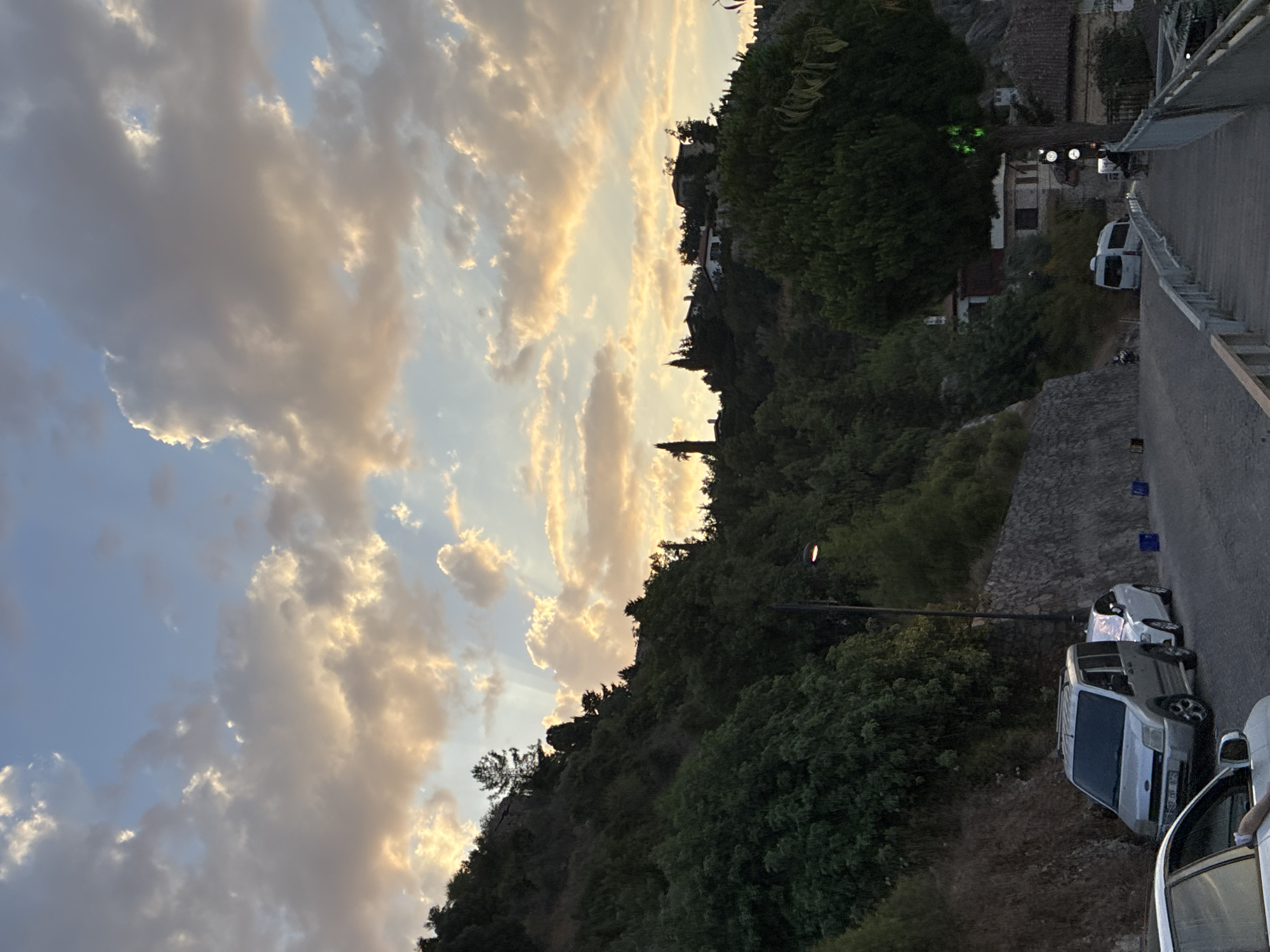 This image captures a scenic view of a hillside road in Alanya, Turkey, likely during dusk. The sky is filled with dramatic clouds tinged with warm orange and yellow hues from the setting sun, with rays of light piercing through. The hillside is densely covered with lush green trees and vegetation. A winding road is visible, with several cars parked along its side, including a silver minivan and a white sedan. Further up the hill, houses and structures are nestled amongst the trees. A pedestrian walkway with a white railing runs alongside the road in the foreground, leading towards a traffic signal and more buildings in the distance. The overall impression is one of a serene, hilly urban landscape bathed in the soft light of twilight.