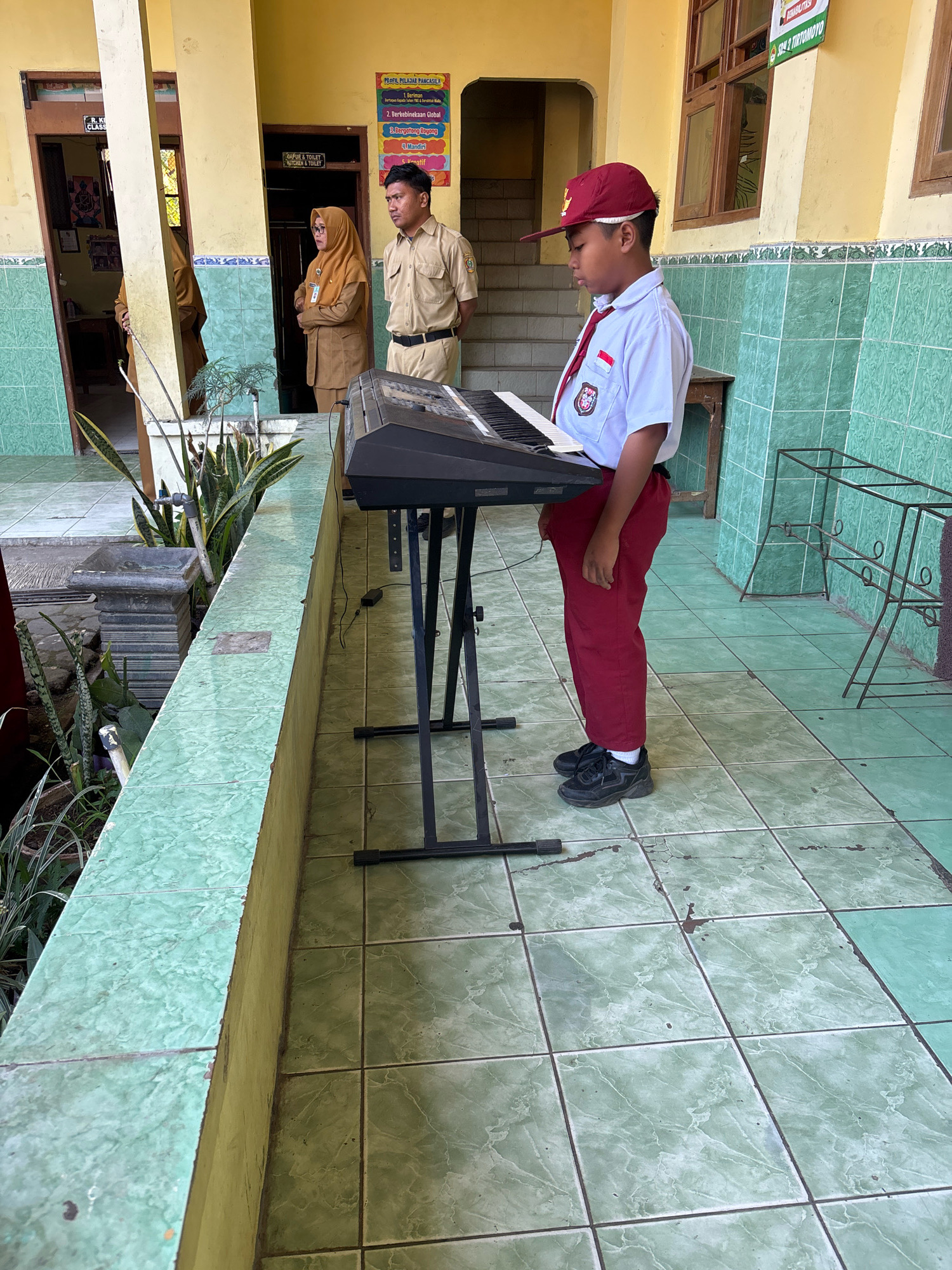 In Tirtomoyo, Indonesia, a young boy in a school uniform, including a red cap and tie, stands before a keyboard on a stand. He is focused on the instrument, his hands positioned as if to play. Behind him, two adults in beige uniforms, likely teachers, observe. One woman wears a headscarf. The setting appears to be an outdoor school corridor, with green tiled walls and a yellow painted upper wall. Plants in pots line a low wall on the left. A bulletin board with colorful text is visible on the wall to the right, indicating school rules or information. The ground is also tiled in green.