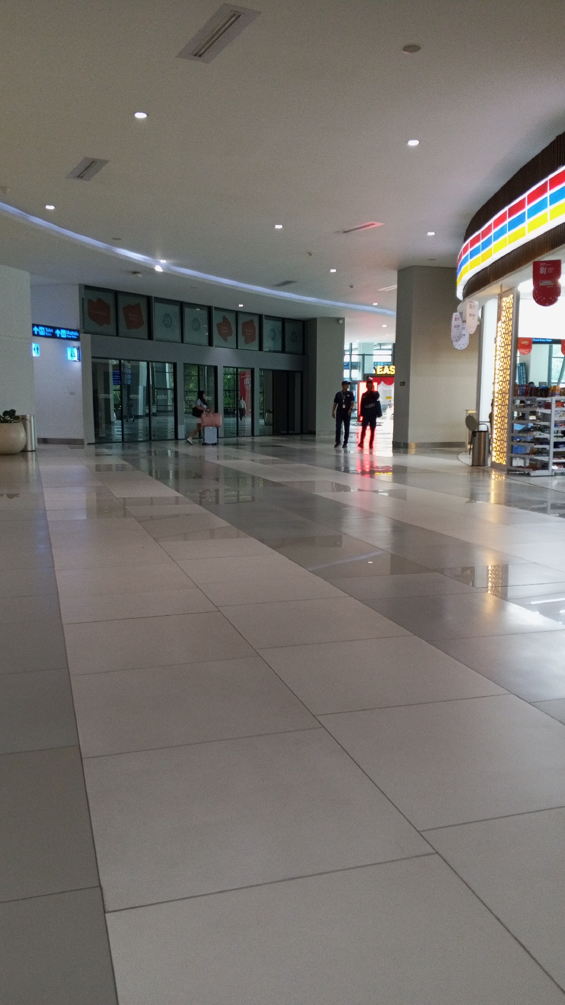 The image depicts the interior of a modern, well-lit shopping mall in Pajang, Indonesia. The floor is covered with large, polished tiles in alternating shades of light grey and beige, creating a pattern of diagonal lines. The ceiling is white with recessed lighting and a curved architectural element with embedded lights.
In the background, a set of glass automatic doors leads into a store. Through the doors, multiple rows of shelves stocked with various items are visible, suggesting a retail environment. Above the entrance, a colorful, curved sign displays the word "EAS" in large yellow letters, with red, blue, and yellow stripes on its border.
Two individuals are walking away from the camera towards the automatic doors. Both are wearing dark colored shirts, and the person on the left appears to be wearing dark pants, while the person on the right is wearing darker pants that reflect the ambient light. A third person is seen walking from left to right in the reflection on the floor near the entrance. Further to the left, a woman is seated on a bench, looking towards the entrance, with a suitcase beside her.
A plant in a large, light-colored pot is situated to the far left, near a waste bin. Blue directional signs with arrows and icons are mounted on a white pillar to the left of the entrance. The overall atmosphere is clean, spacious, and orderly, characteristic of a contemporary commercial space.