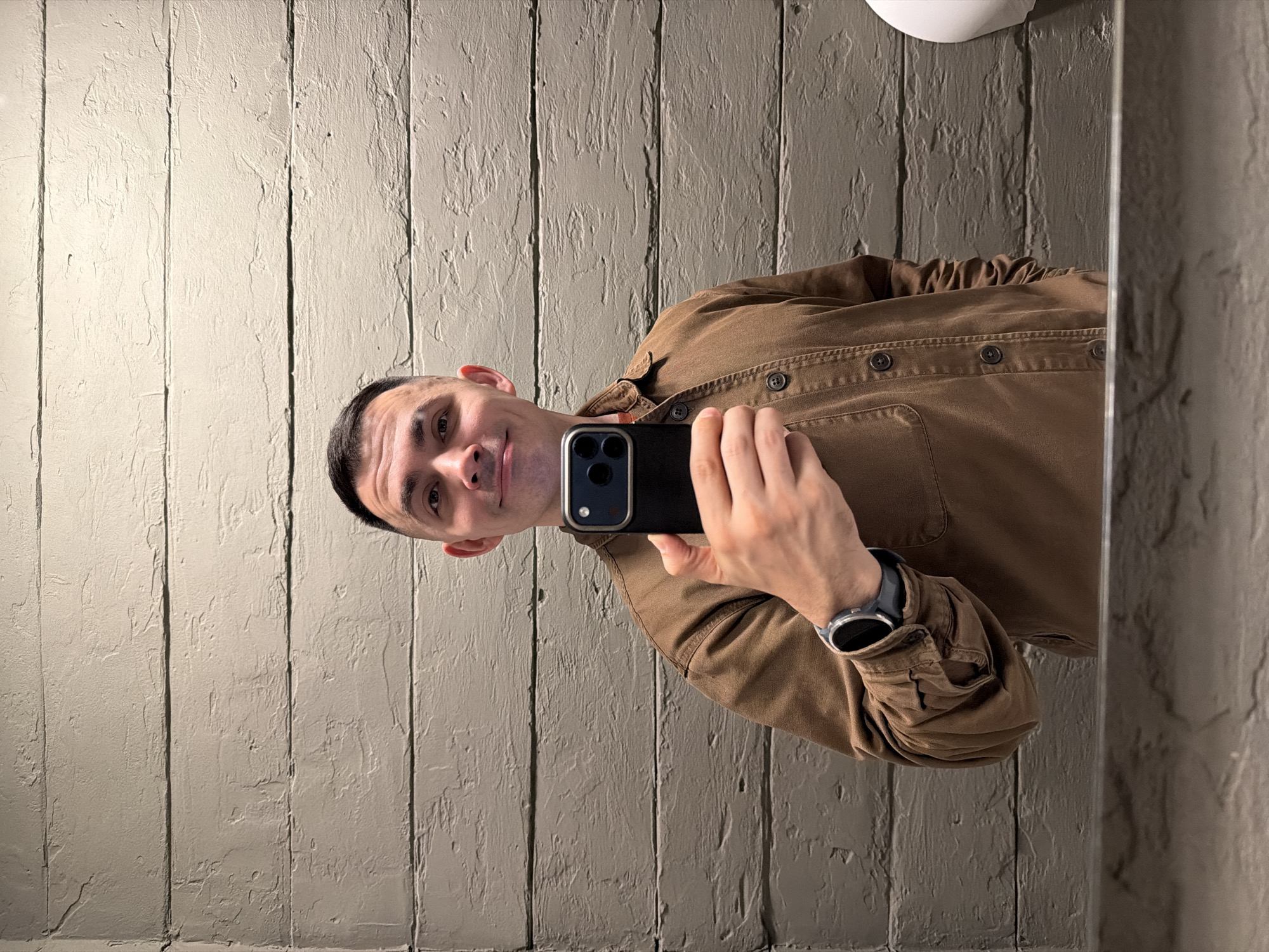 A man is captured in a mirror selfie, looking directly at the camera with a gentle smile. He has short, dark hair and is wearing a collared, long-sleeved brown or olive-brown shirt or jacket. On his left wrist, he wears a dark-colored smartwatch. He holds a black smartphone, likely an iPhone, with both hands; its camera module with two lenses is visible.
The man is positioned against a textured wall, which appears to be composed of vertical panels with a rough, stucco-like finish in a light, desaturated greenish-gray or beige hue. The lighting seems to be artificial, highlighting the wall's uneven surface. A small white object, possibly a light fixture or part of a sink, is visible at the top edge of the frame. The overall atmosphere is casual and direct. The image was taken in Izhevsk, Russia.