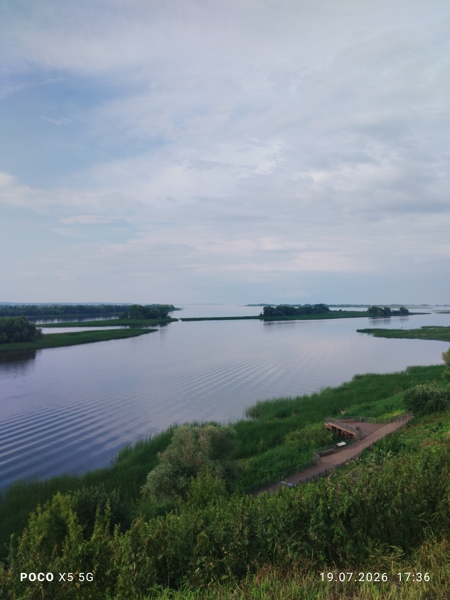 An aerial, wide-angle shot captures a serene landscape featuring a wide, calm river or lake. The water's surface is disturbed by a series of parallel ripples, suggesting movement from a passing boat or wind. Lush green reeds and grasses line the banks, creating a vibrant contrast with the muted tones of the water and sky. Small, tree-covered islands dot the expanse of water, adding depth and visual interest to the scene. In the foreground, a verdant slope descends towards the water, adorned with dense foliage and vegetation. A wooden walkway with railings and a viewing platform is integrated into the slope, providing access to the scenic overlook. The sky above is overcast, with soft, diffused light casting a gentle glow over the entire vista. The timestamp "19.07.2026 17:36" and the text "POCO X5 5G" are visible in the lower corners of the image, indicating the date, time, and device used for capture. This scene is likely located near the city of Bolgar, Russia, given the prominent Volga River and its surrounding natural beauty.