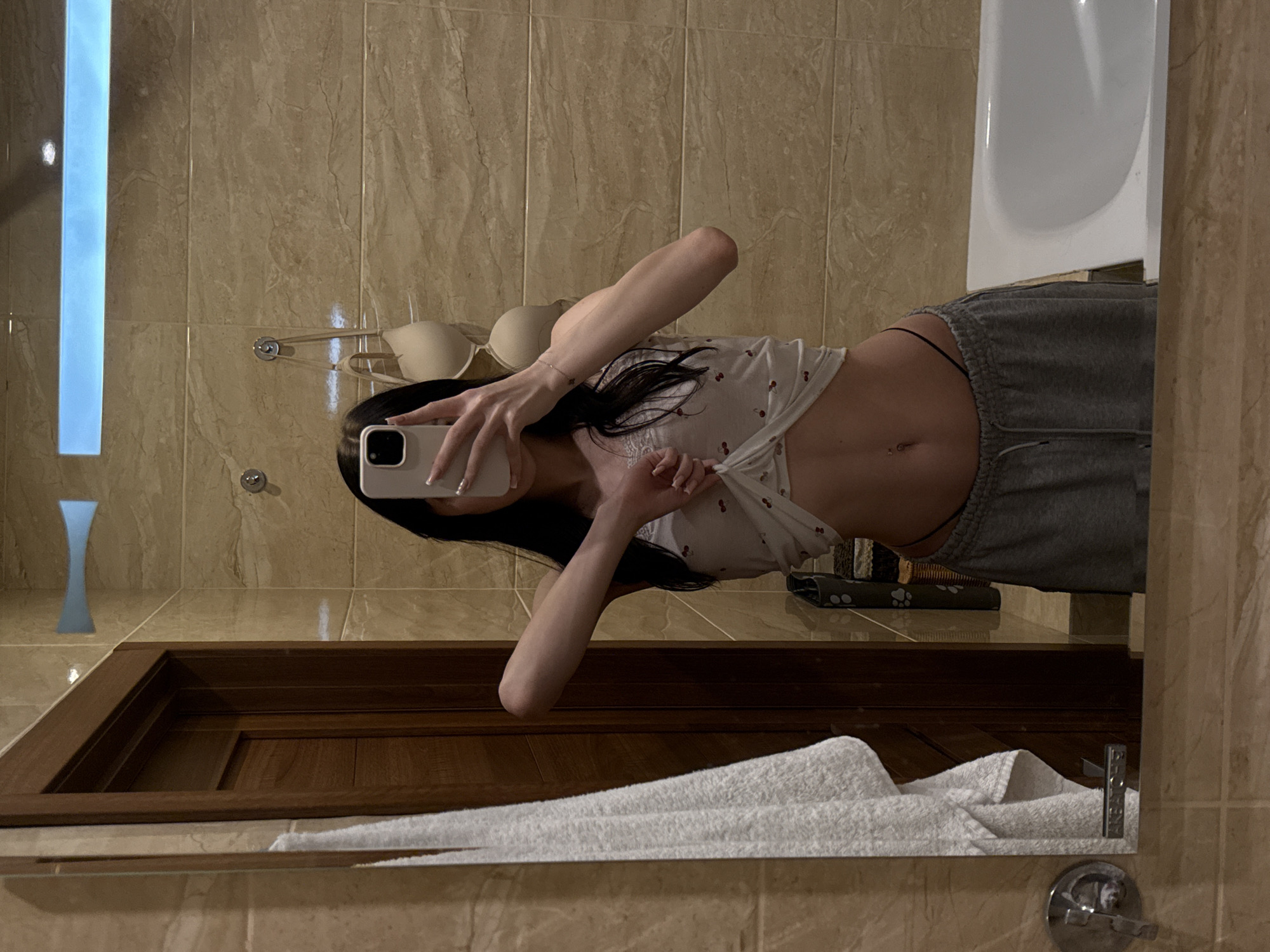 A young woman is taking a mirror selfie in a bathroom. She is wearing a white crop top with red cherry patterns and grey sweatpants. Her hair is dark and falls past her shoulders. She is holding a light-colored smartphone in her right hand, capturing her reflection. The bathroom has tiled walls and a white bathtub is visible on the right side of the frame. A towel hangs on the left, and a wooden door is reflected in the mirror. A bra is hanging on the wall behind her. The scene appears to be in a private residence.