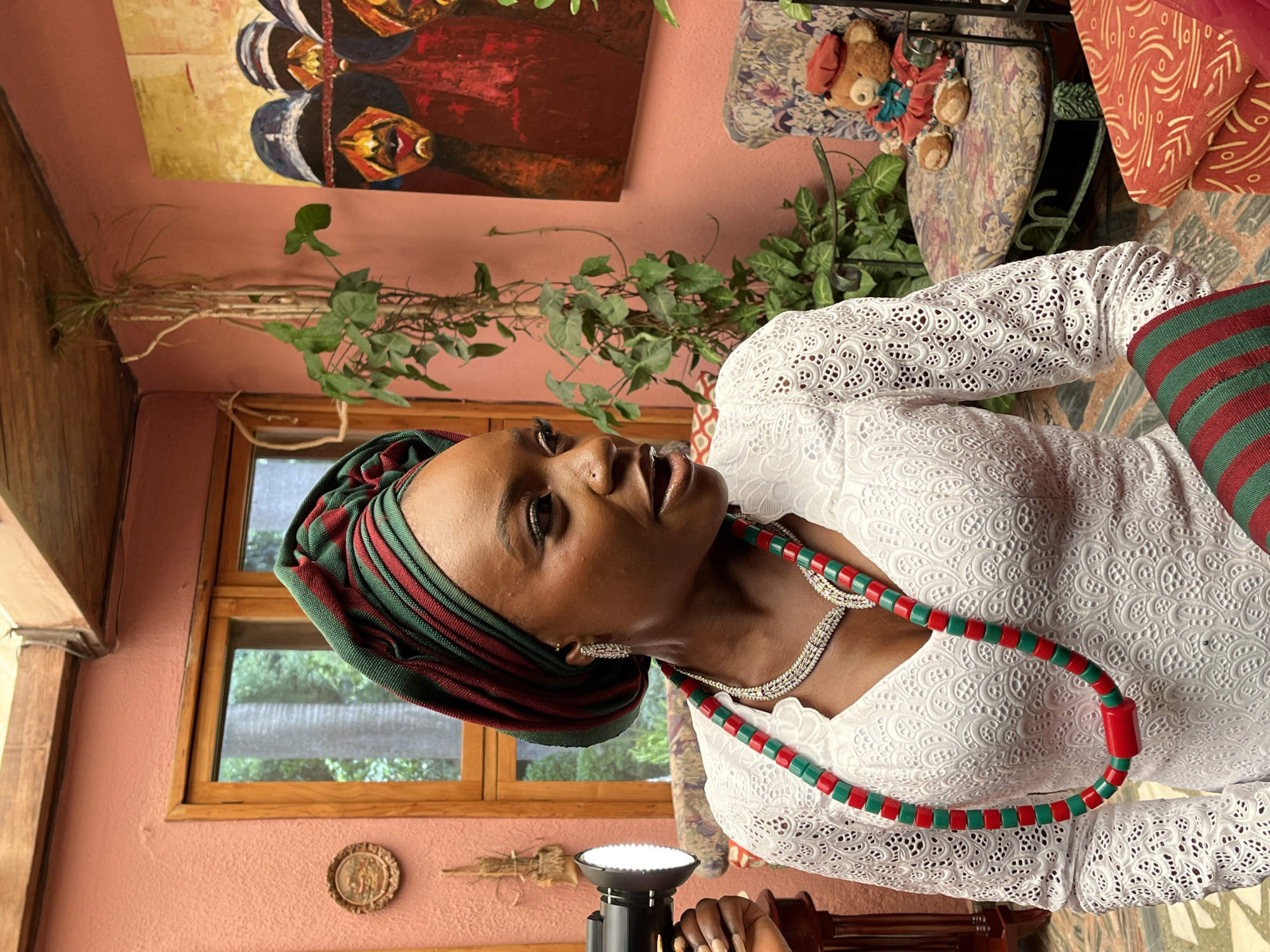 A medium shot shows a Black woman wearing a white, intricately patterned lace dress with a v-neckline and long sleeves. She has a dark green and red striped headwrap, a matching beaded necklace with red cylindrical beads and green square beads, and a diamond-encrusted necklace. Her hair is pulled back. She is looking to the right, with her mouth slightly open. To her left, a hand is holding a black spotlight with a white light.
The background includes a salmon-pink wall, a wooden-framed window with greenery visible outside, a hanging vine plant, and a painting on the wall depicting three figures wearing hats and red and black clothing. To the right, there is a chair with a floral pattern, a teddy bear wearing a red beret and a red outfit, and an orange cushion with a white pattern. The floor has a tiled pattern. The scene appears to be an interior space, possibly a studio or a decorated room. The image is cropped to focus on the woman and her immediate surroundings. The location is identified as Bar Jirgi Summa, Nigeria.