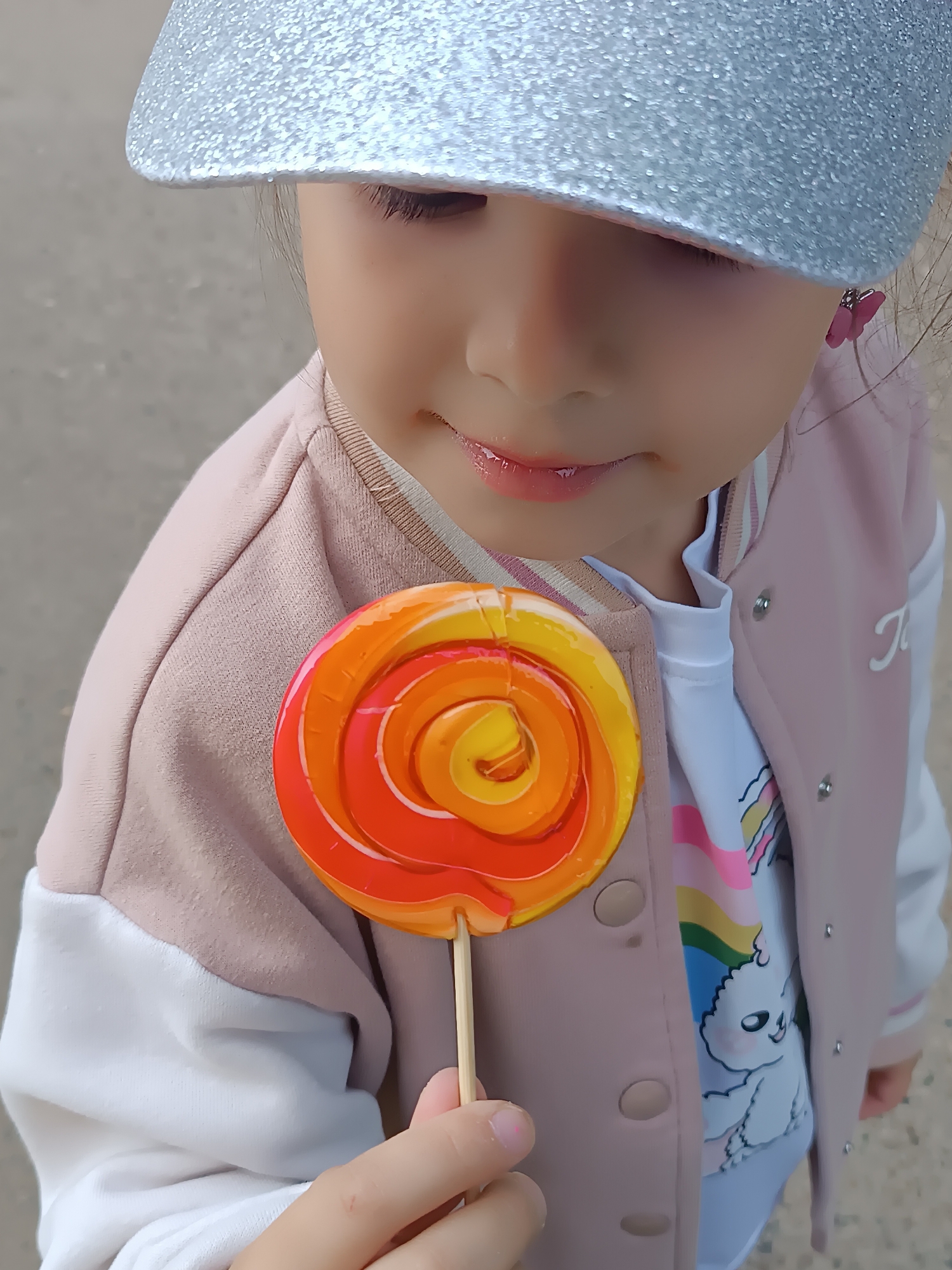 A close-up, eye-level shot captures a young child holding a large, vibrantly colored swirl lollipop. The child, likely a girl, is shown from the chest upwards, with her face partially obscured by a sparkly silver visor or hat, which covers her eyes. Her lower face, including her nose, mouth, and chin, is visible, revealing slightly parted, glossy lips with a hint of a gentle smile. She appears to be looking down at the lollipop with focused attention.
The lollipop itself is a prominent feature, circular in shape with a striking spiral pattern of orange, yellow, and red hues, held by a light wooden stick in the child's right hand.
The child is dressed in a light pink and white varsity-style jacket or cardigan with button fasteners, worn over a white t-shirt. The t-shirt features a colorful graphic, including what appears to be a rainbow design and a cartoon-like white fluffy animal, possibly a sheep or bear. A small pink butterfly earring is visible in her right ear. The background is out of focus, appearing as a soft, neutral gray-beige, suggesting an outdoor or brightly lit indoor setting. The overall lighting is bright, indicating daytime. There is a faint letter or symbol, possibly "T", on the white part of the jacket on the right side of the frame.