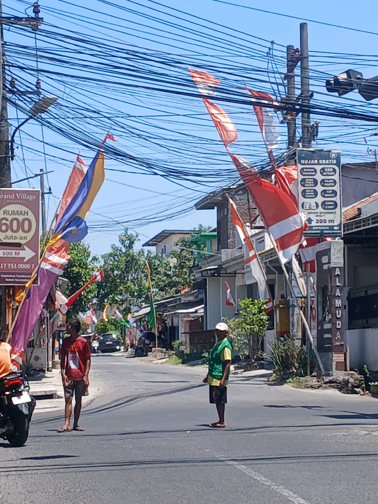 In Surabaya, Indonesia, two men stand on an asphalt road during the daytime. The man on the left, wearing a red t-shirt and patterned shorts, stands facing right. To his right, another man, wearing a yellow shirt and black shorts, stands facing left. A motorcycle is parked to the left, and other motorcycles are visible further down the road, with a black car also in motion. The street is lined with buildings and trees. Numerous Indonesian flags and colorful banners are strung across power lines and poles, creating a festive atmosphere. A sign is visible on a pole with text that includes "HUJAN GRATIS" and "200 m". The sky is clear and blue.