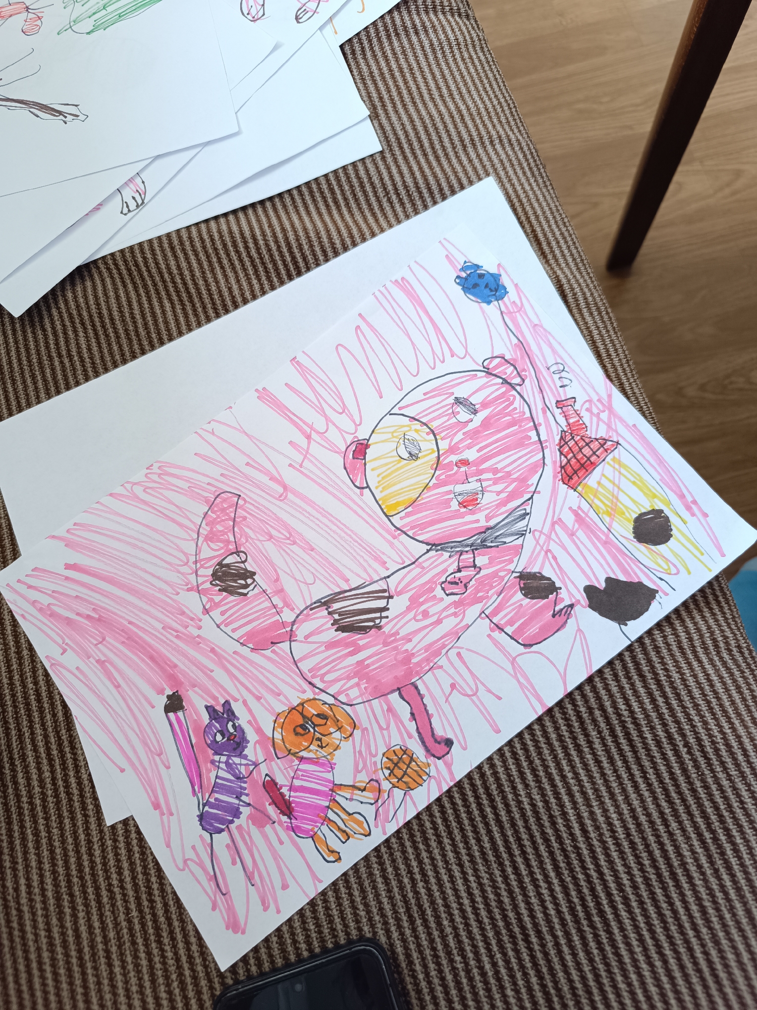 A child's drawing lies on a woven brown surface. The drawing depicts a pink bear-like creature with a yellow face and black collar holding a bottle rocket with brown spots. A blue fish floats above the bear. To the left of the bear, a purple cat and orange dog, both striped, stand on the ground. Other drawings are stacked in the upper left corner. The scene is indoors. The city and location are unknown.