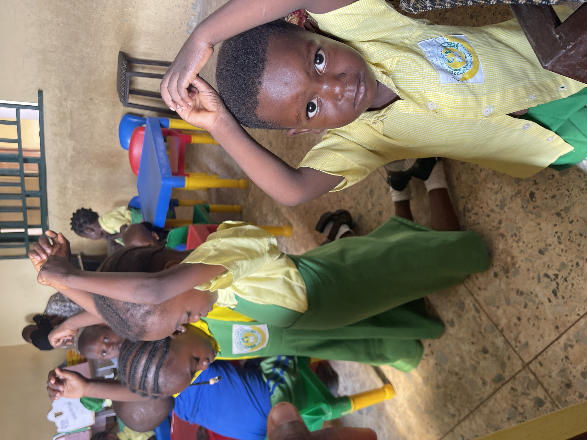 The image captures a vibrant indoor scene, likely a classroom or learning environment, filled with young Black children. The primary subject is a young child, possibly a boy, in the foreground who looks directly at the viewer with a neutral, serious expression. He wears a yellow checkered short-sleeved shirt and green pants, with his arms bent and hands clasped over his head. A circular emblem, primarily white with green and yellow designs, is visible on his left chest pocket, likely a school logo.
Several other children are present in the midground and background, mostly seen from behind or the side. They are uniformly dressed in similar yellow checkered shirts and green skirts or pants, suggesting school attire. One child in the midground wears a blue shirt with a green skirt. Many children have braided hairstyles. They appear to be seated or standing around small, colorful plastic tables and chairs (blue, red, and yellow), characteristic of an early childhood education setting.
The setting features a light-colored tiled floor and plain walls. A window with horizontal bars is partially visible on the left. The lighting suggests it is daytime. A dark wooden stool or chair is in the background. The children seem engaged in an activity, though specific details are not clear. A partial view of an adult's hand is visible in the lower right corner, possibly indicating an adult present in the room or taking the photograph. The visible text on the children's emblems appears to be a logo, but specific letters or words are not clearly legible. The scene depicts a typical school day in Akwanga, Nigeria, given the provided context.