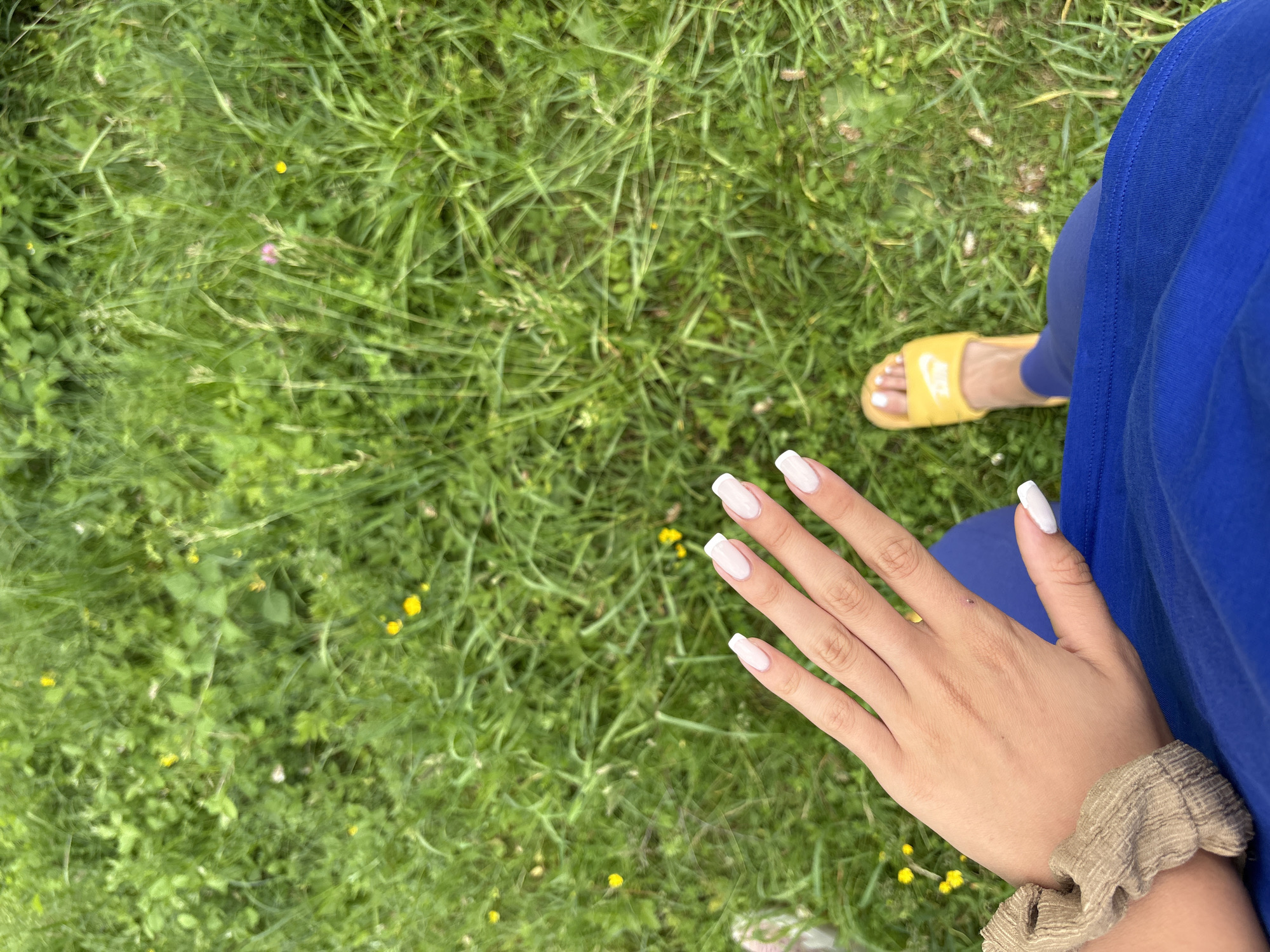 A person's hand with white-tipped nails is held up against a background of green grass with small yellow flowers. The person is wearing a blue outfit and a yellow slide sandal.