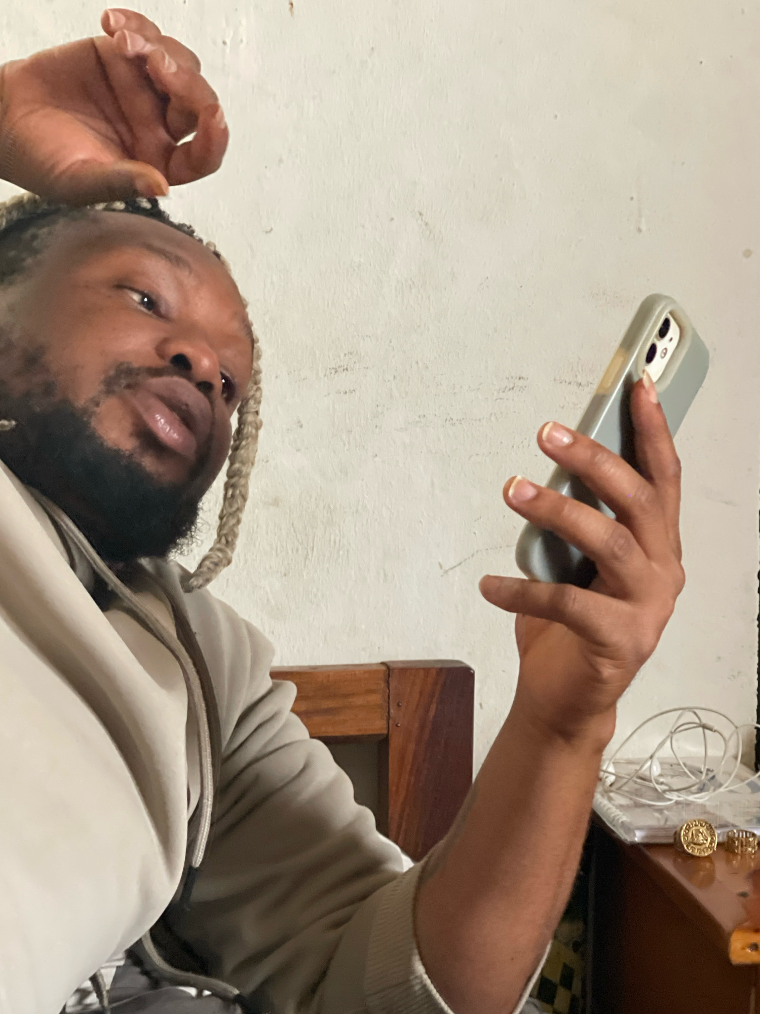 In a close-up, indoor shot, a person with braided, light-colored hair and a beard holds a light gray smartphone in their right hand, which is positioned in the foreground. The person's left hand is raised near their head, with the index finger and thumb forming a circle. They are wearing a light beige hooded sweatshirt. The background consists of a textured, off-white wall and a wooden furniture item. On a surface to the right, a white cord is coiled next to a stack of thin books or papers, with two gold rings placed on top. This scene is consistent with a personal space, potentially a bedroom or living area within a home in Jos, Nigeria.