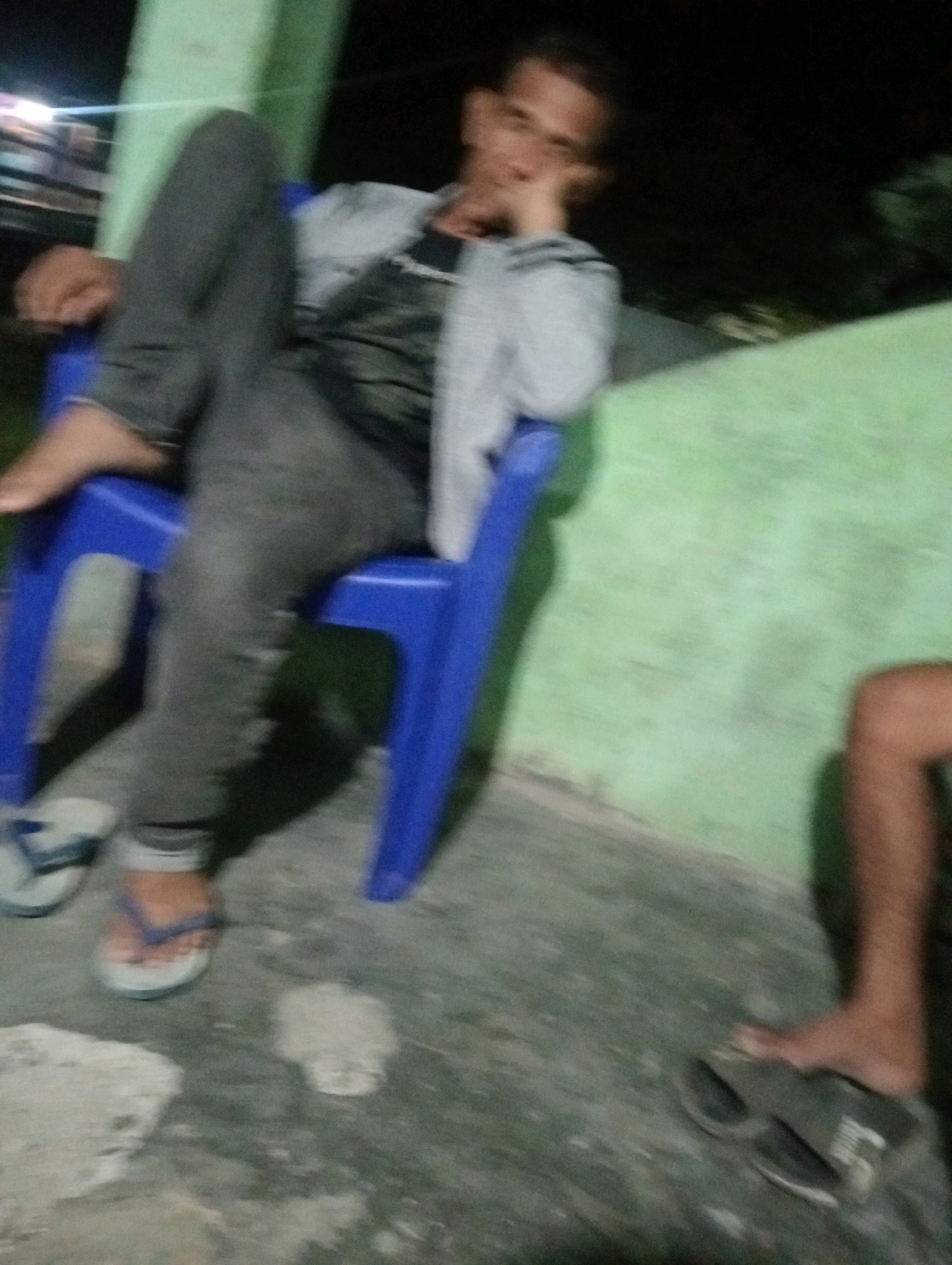 A blurry, low-light image captures a person sitting on a blue plastic chair in what appears to be an outdoor area with a green wall in the background. The individual is wearing gray pants and a light-colored jacket over a dark shirt. Their left leg is extended, and their right hand is resting on their chin, suggesting a relaxed or contemplative posture. In the foreground, a pair of white flip-flops are visible near the person's feet. Another person's bare leg and foot, wearing dark sandals, is partially visible on the right side of the frame. The ground is concrete, with some debris. The overall impression is of a casual, possibly evening scene in an urban or semi-urban setting in Tanjung Pandan, Indonesia, given the green painted wall, common in such locations.