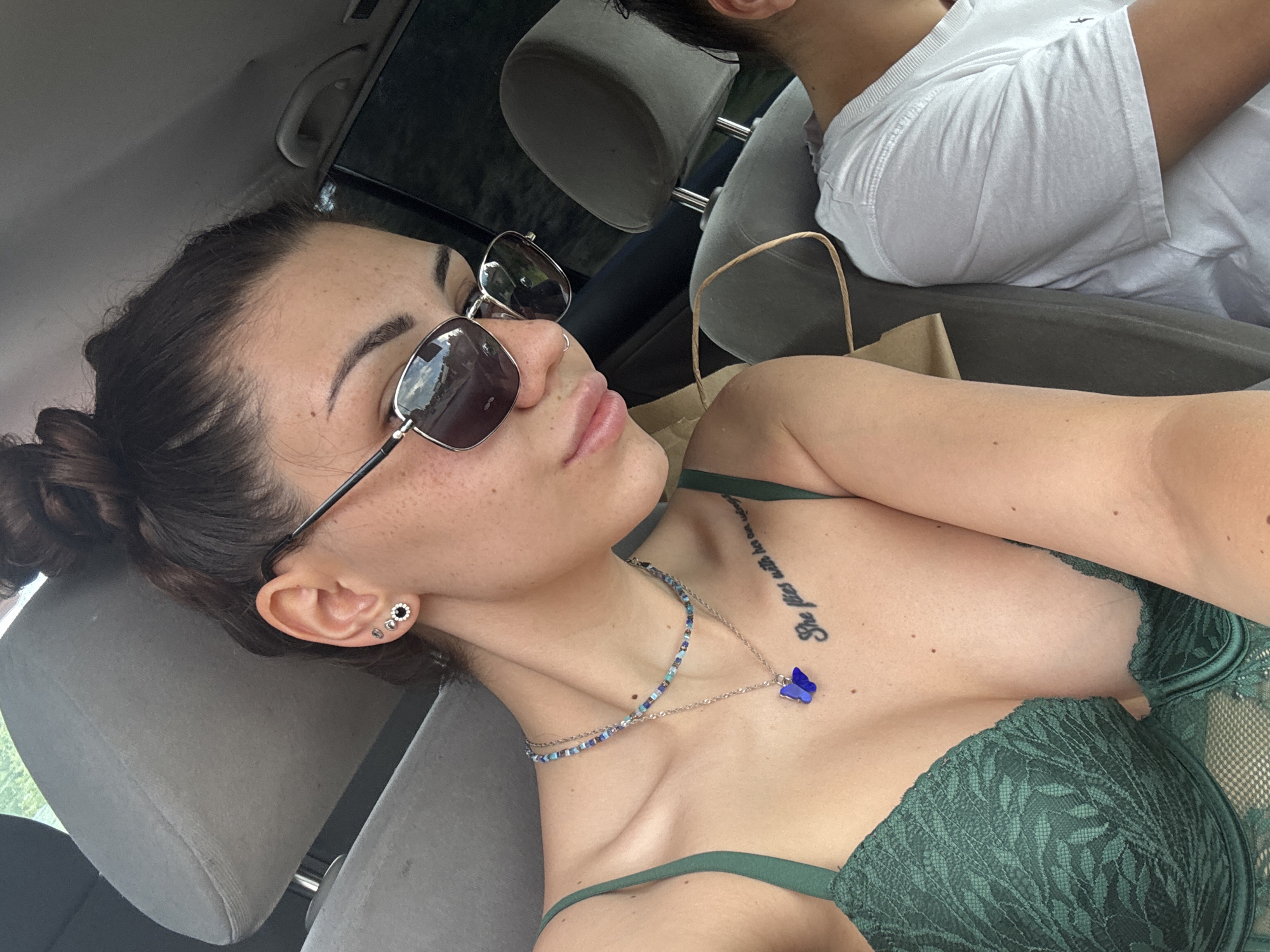 The image captures a woman and a partially visible man inside a car, likely in Gorna Malina, Bulgaria, during daytime. The primary subject is a woman, positioned in the foreground as if in the driver's seat, with her head tilted back and eyes closed or looking downwards, creating a relaxed expression. She has dark hair pulled back in a bun and visible freckles across her face and chest.
She is wearing dark sunglasses, a silver nose ring, and multiple ear piercings, including a prominent silver stud with a black center. Her attire consists of a dark green lace bra with a leaf pattern. She adorns two necklaces: one beaded in blue and silver, and another silver chain featuring a distinctive blue butterfly pendant. A tattoo in black script is visible on her left collarbone/chest area, reading "She flies with her own wings".
In the background, a man is partially seen in the passenger seat, facing away from the camera. He wears a white t-shirt. A brown paper bag is visible next to him. The car's interior features grey headrests. Reflections in the woman's sunglasses suggest daylight conditions, possibly with greenery and sky outside the vehicle. The scene conveys a moment of rest or travel.
