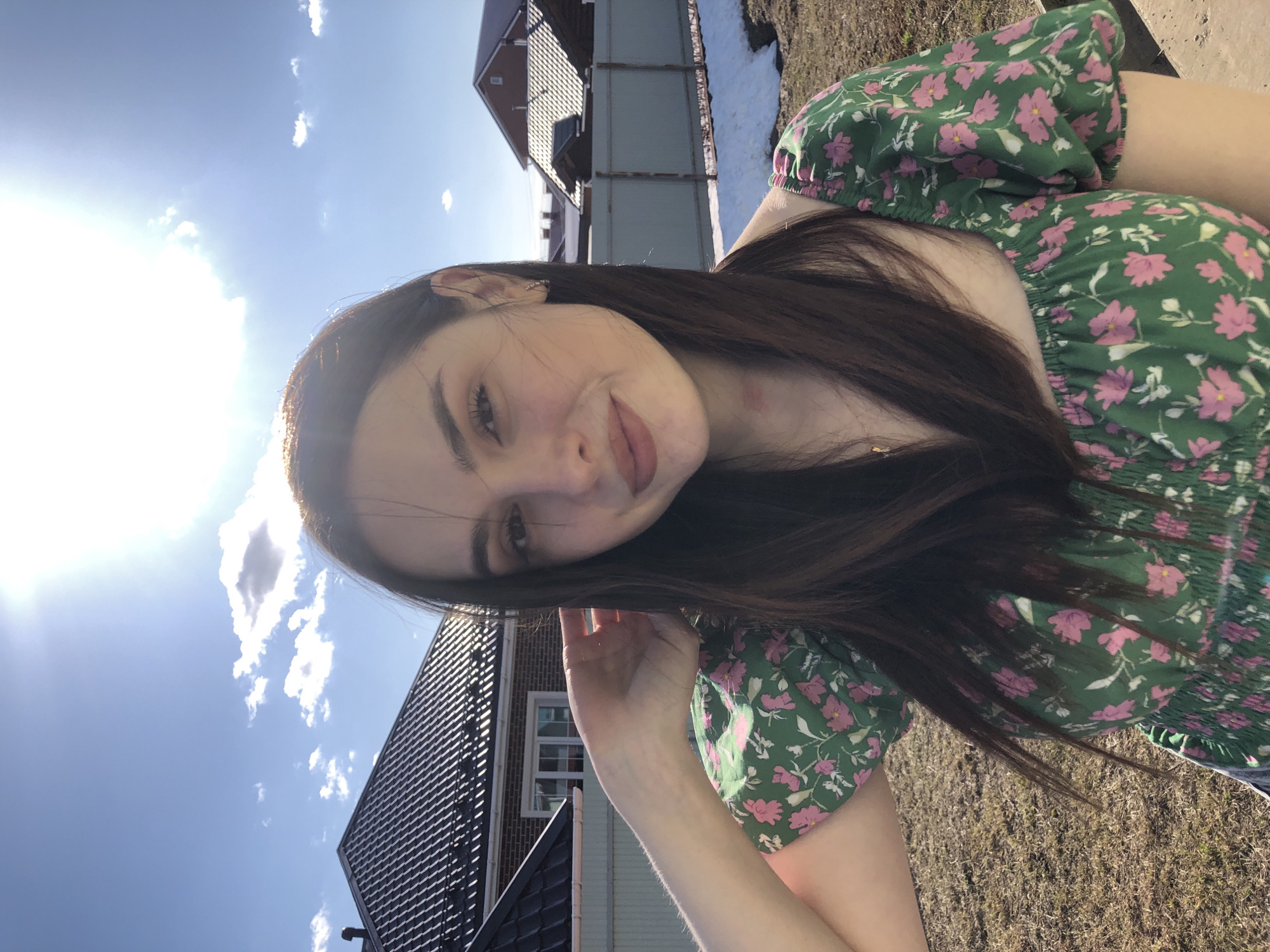 A young woman poses for a selfie outdoors on a sunny day. She has long, dark brown hair and is wearing a green dress with pink floral patterns. She is smiling slightly, and her left hand is near her face. In the background, there are houses with black roofs and some melting snow on the ground. The sky is blue with scattered clouds. The location appears to be in a residential area, and the overall lighting suggests it is daytime.