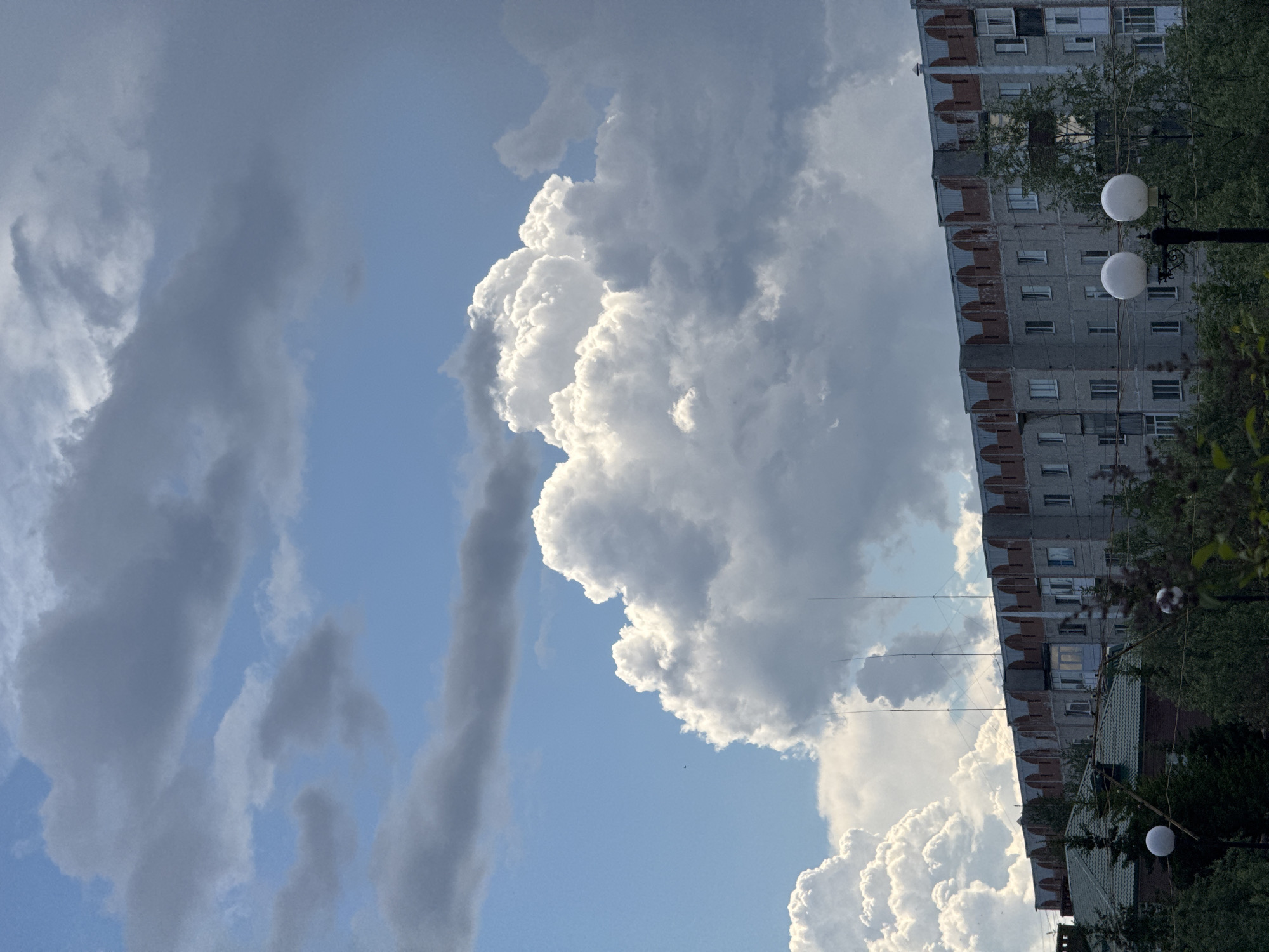 This is a vertical, eye-level photograph taken from a slightly elevated position, looking towards a multi-story apartment building in Megion, Russia. The sky dominates the upper half of the frame, filled with dramatic, billowing cumulus clouds illuminated by sunlight, interspersed with darker, more ominous storm clouds. A small, dark bird is visible in flight against the blue sky to the left of the largest cloud formation.
The apartment building, situated in the middle ground, appears to be a typical Soviet-era block with a grey facade, numerous windows, and a decorative cornice along the roofline featuring arched elements. Three tall, thin antennas protrude from the roof of the building. In front of the building, a line of green trees and bushes obscures the lower levels, with the top of another building with a green roof visible to the far left. Two white, globe-shaped streetlights are positioned in the foreground, slightly to the right of center, with one being partially obscured by foliage. The overall impression is of a residential area under a dynamic, possibly stormy sky.