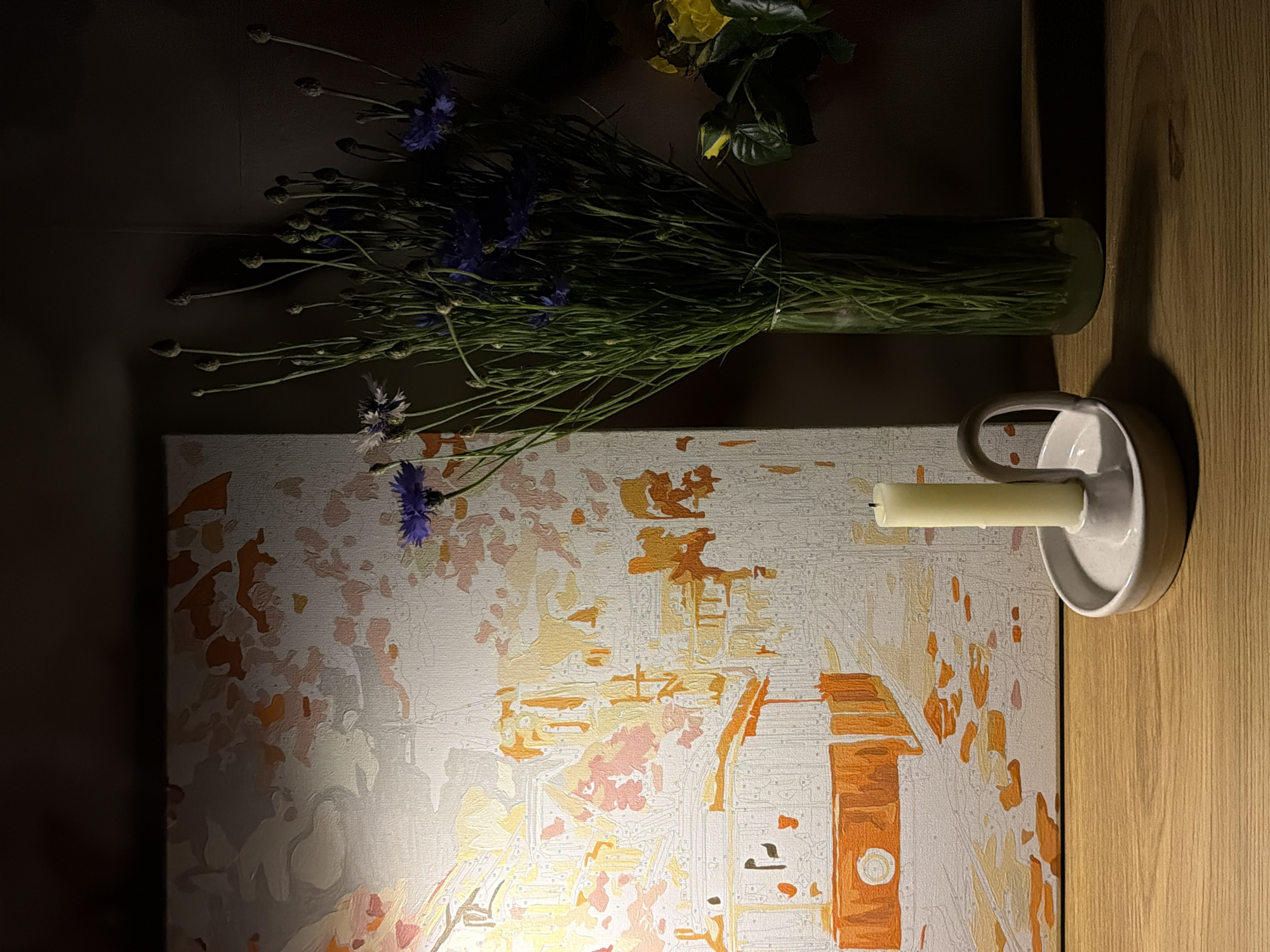 A still life composition features a partially completed paint-by-numbers canvas depicting a building in shades of orange, white, and pale pink. Atop the canvas sits a cylindrical glass vase filled with a dense arrangement of blue cornflowers and green foliage. A white ceramic candle holder containing an unlit pale yellow candle is positioned to the right of the canvas. The scene is illuminated by a warm light source, likely artificial, casting shadows and highlighting textures. The canvas rests on a dark surface with a wooden edge visible on the right side of the frame. The exact location and city are not identifiable from the image.