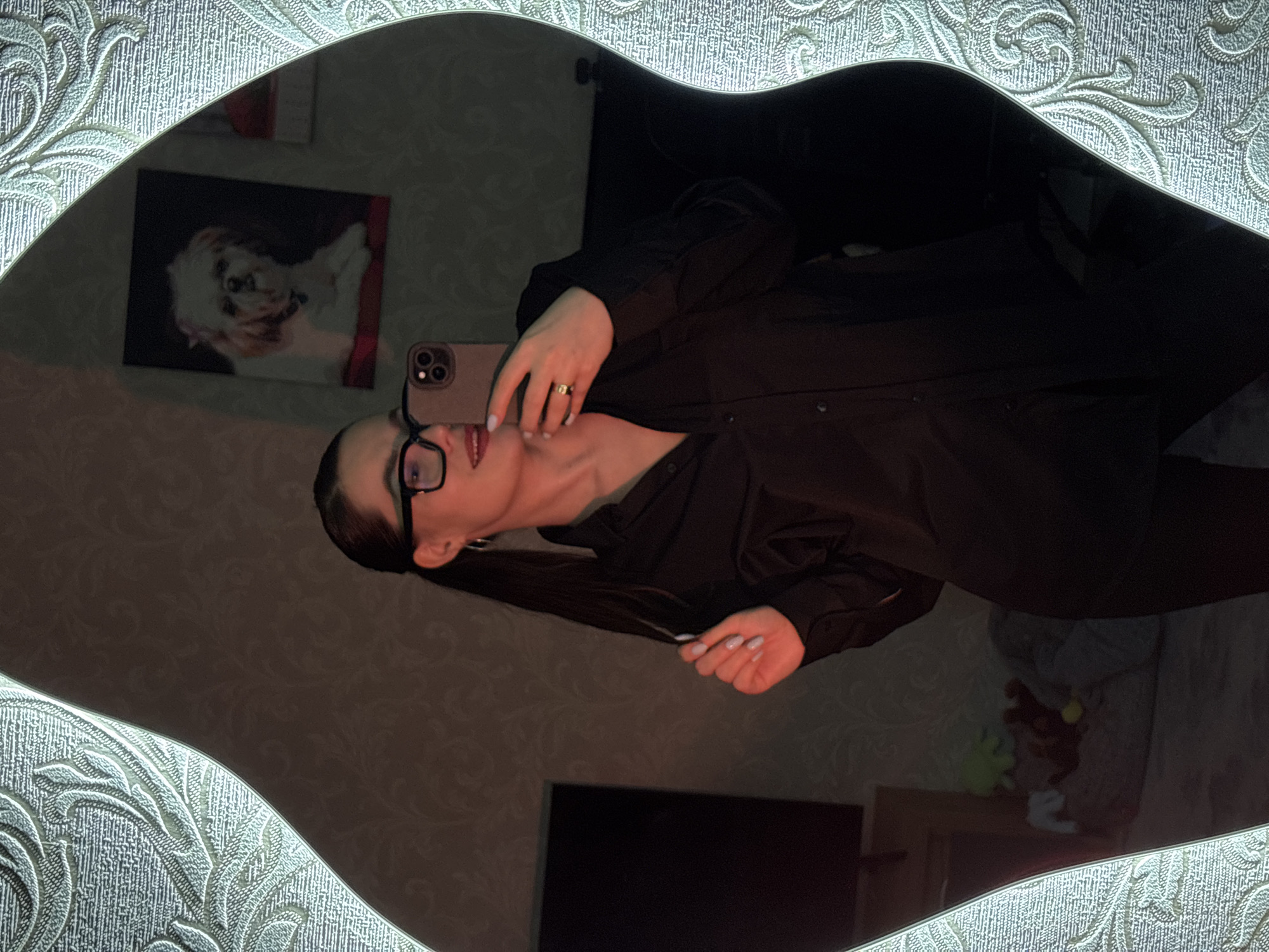 A woman is reflected in a mirror with an ornate white frame. The woman has dark hair styled in pigtails, glasses, and is wearing a dark button-down shirt. She is holding a smartphone in her hand, presumably taking the picture. The reflection in the mirror shows part of a room with patterned wallpaper. On the wall, a picture of a dog can be seen. A television and some toys are visible on a shelf below. While this gives a general impression of the setting, information about whether the photograph was taken in Chaikovsky, Russia is not available.