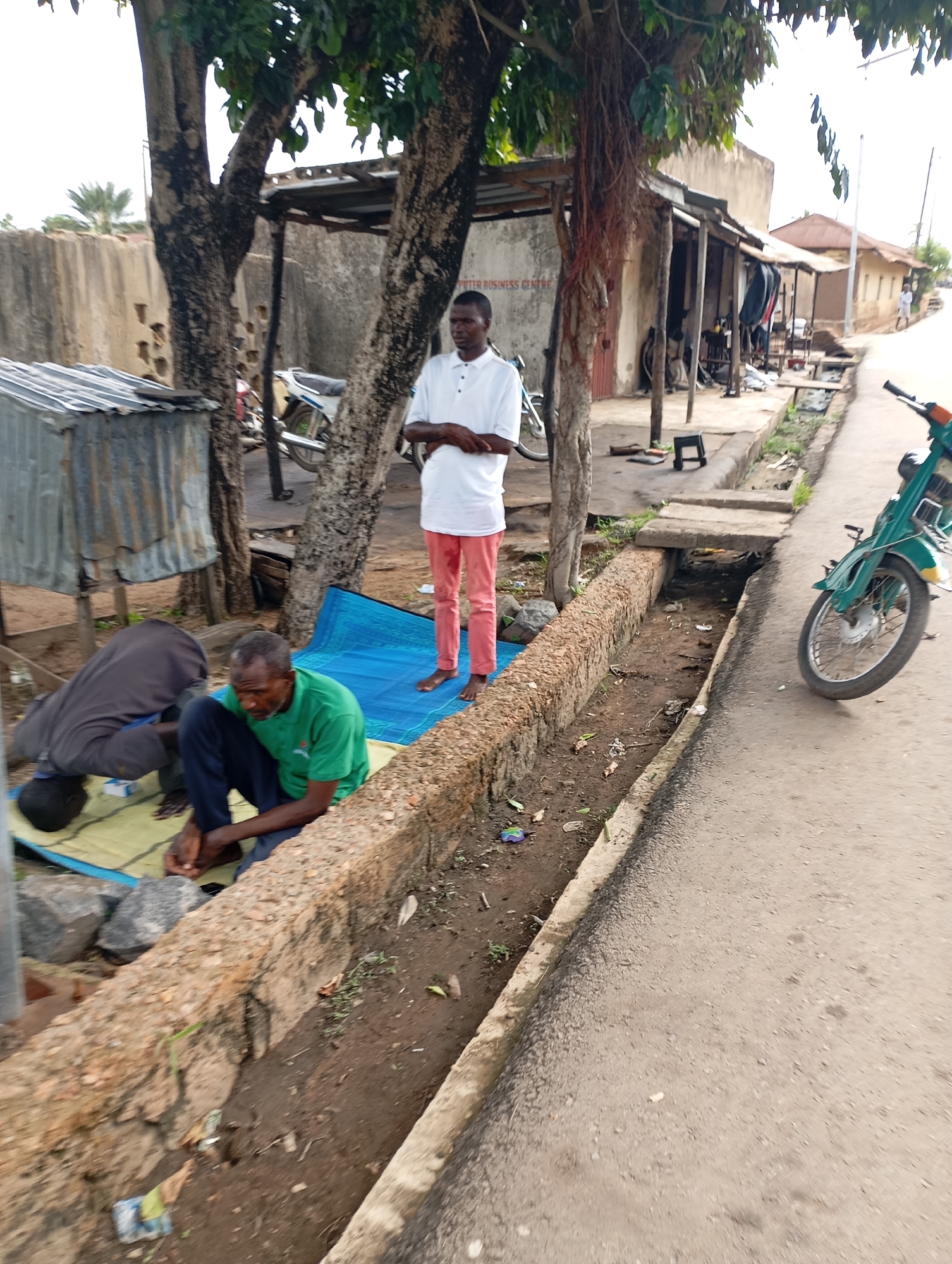 A medium shot captures an outdoor scene in Shendam, Nigeria, on a street beside a rudimentary open drainage ditch. In the foreground, two men are visible on a blue mat. One man, wearing a green t-shirt and dark pants, is seated and appears to be interacting with something in his hands. Behind him, another man is in a prayer position, with his head lowered towards the ground. To the right, a portion of a green motorcycle is parked on the asphalt road. Further back, a man in a white t-shirt and pink pants stands near some trees and a building. The building has a corrugated metal roof and appears to be a small shop or business. The environment suggests a less developed urban area, with a dirt and debris-filled drainage ditch and simple structures. The sky is overcast.