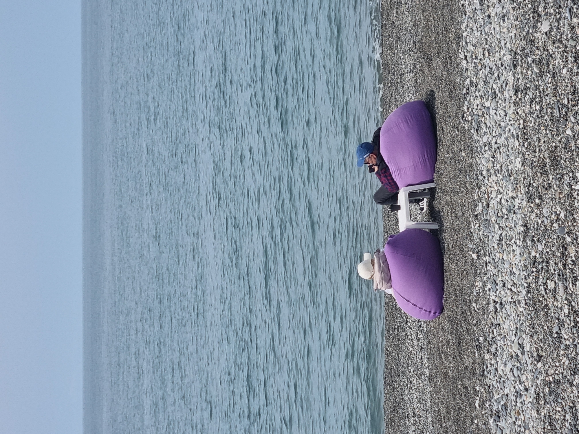This image captures a tranquil beach scene, likely in Sochi, Russia, on a bright and clear day. The setting features a vast expanse of calm, light blue sea stretching to a hazy horizon under an equally clear sky. In the foreground and midground, a wide pebbly beach, composed of various shades of grey stones, lines the water's edge.
Two individuals are relaxing on the beach. Each person is seated comfortably on a large, vibrant purple beanbag chair, accompanied by a small, minimalist white stool or table placed in front. The person on the left wears a blue baseball cap and a plaid shirt, appearing to be engrossed with a mobile phone they are holding. The second person, positioned further to the right, is wearing a light-colored, possibly white or cream, hat or hooded top. They are also seated in a relaxed posture on their purple beanbag.
The sea shows gentle ripples on its surface, reflecting the soft light of the day. The overall atmosphere suggests a peaceful moment of leisure by the coast, with both individuals enjoying the pleasant weather and serene surroundings. There is no visible text in the image.
